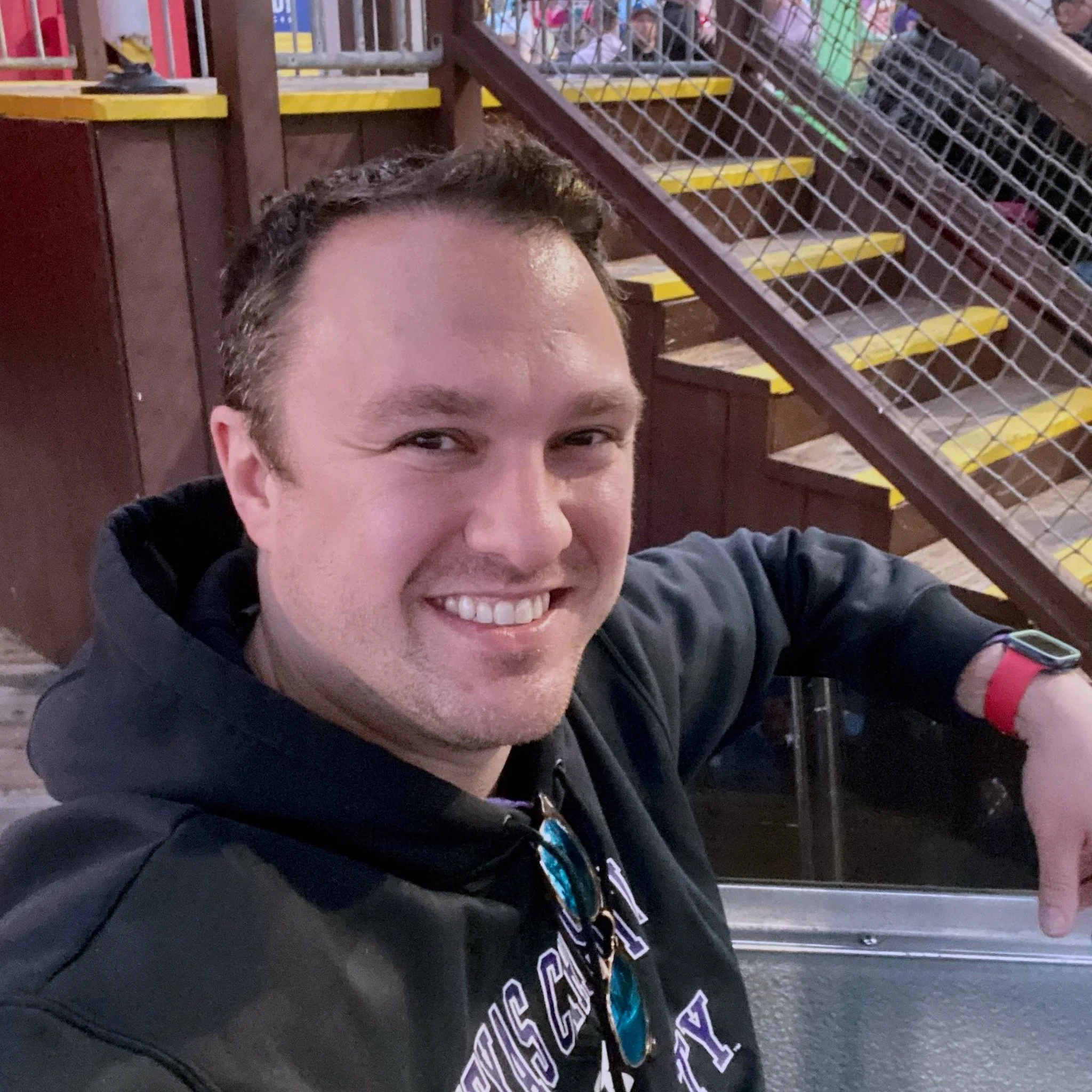 A man smiling and pointing at something in an indoor amusement park or arcade setting with stairs and colorful equipment in the background.