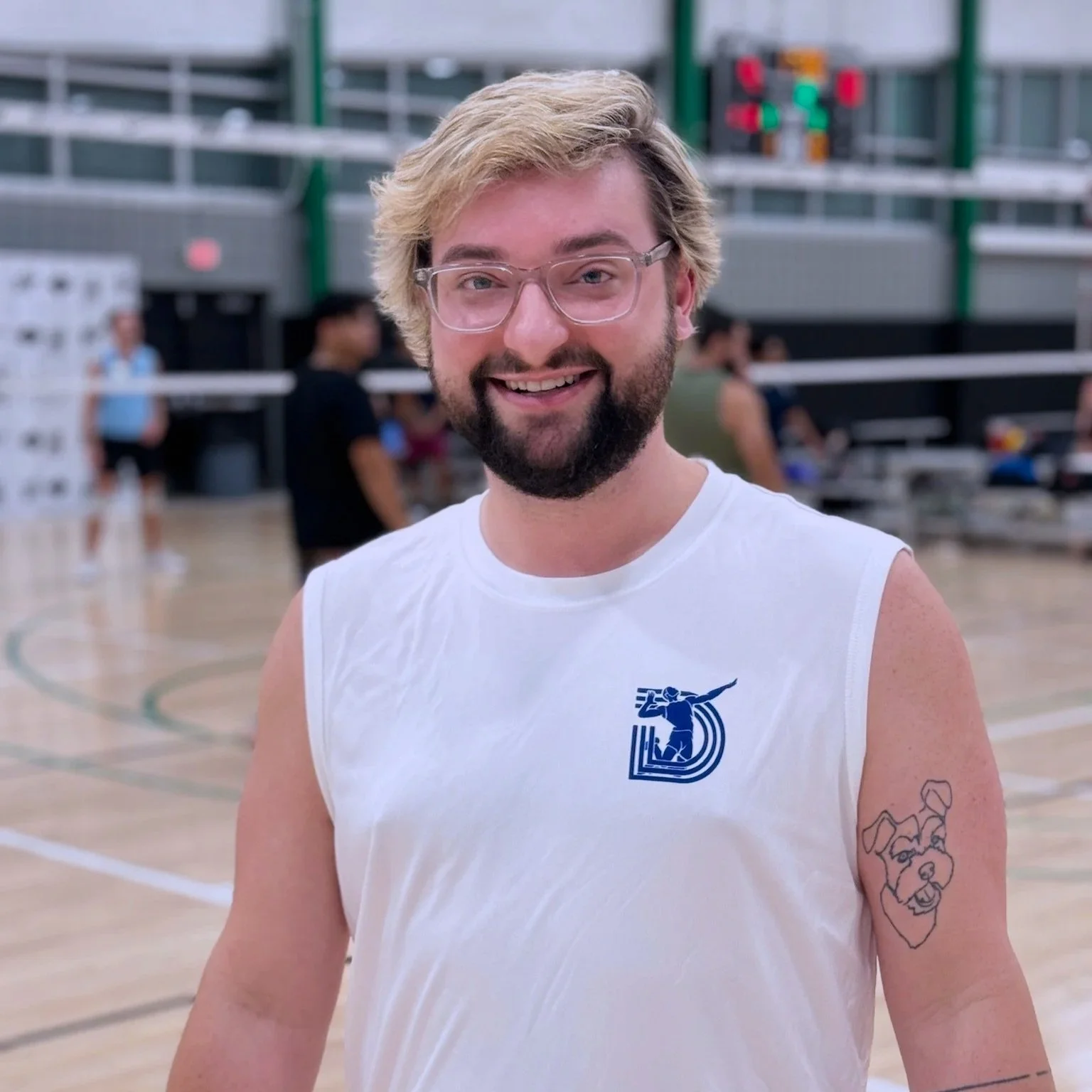 A smiling man with glasses and a beard wearing a white sleeveless sports shirt, standing on a basketball court. There are people and seating in the background.