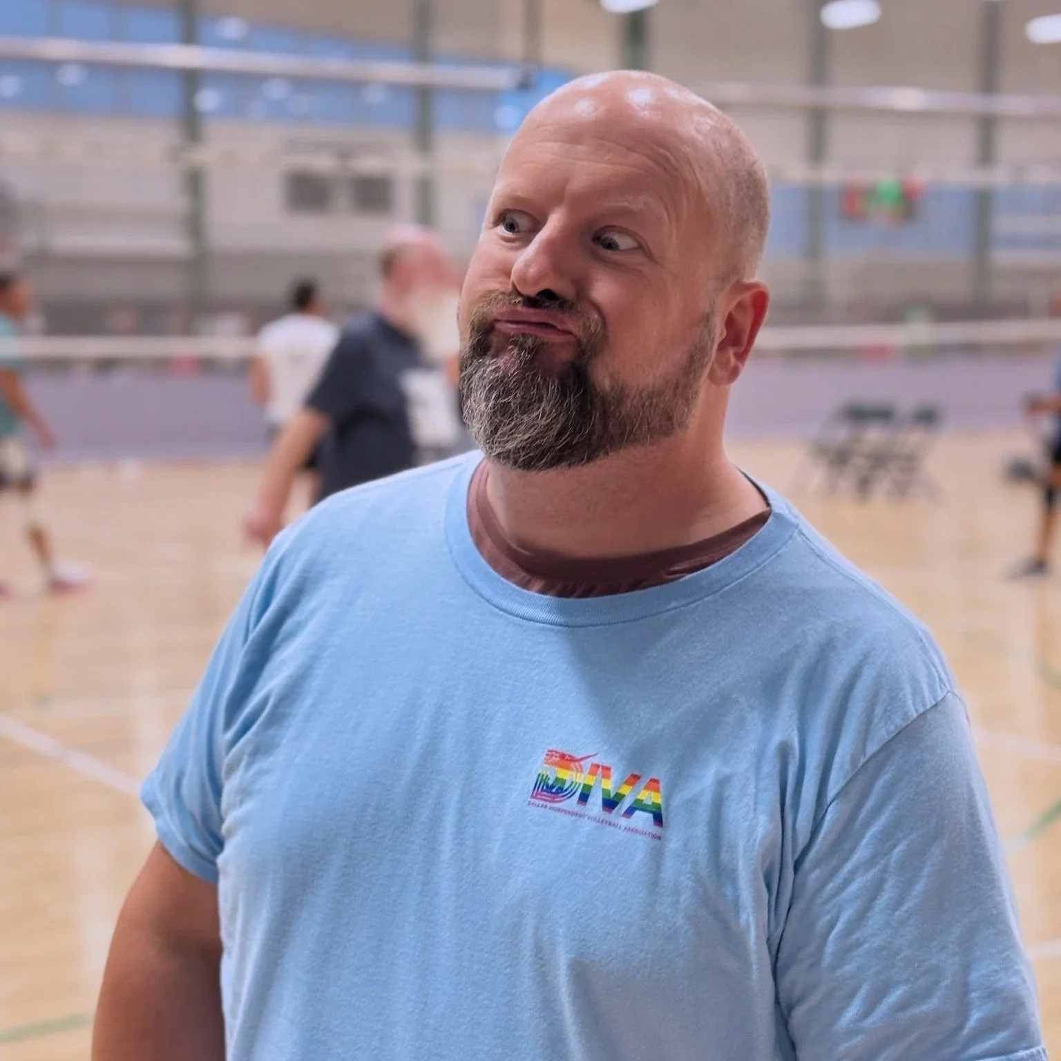 A man with a shaved head and a beard, wearing a light blue T-shirt with a rainbow-colored logo, standing in an indoor sports facility with people and a basketball court in the background.