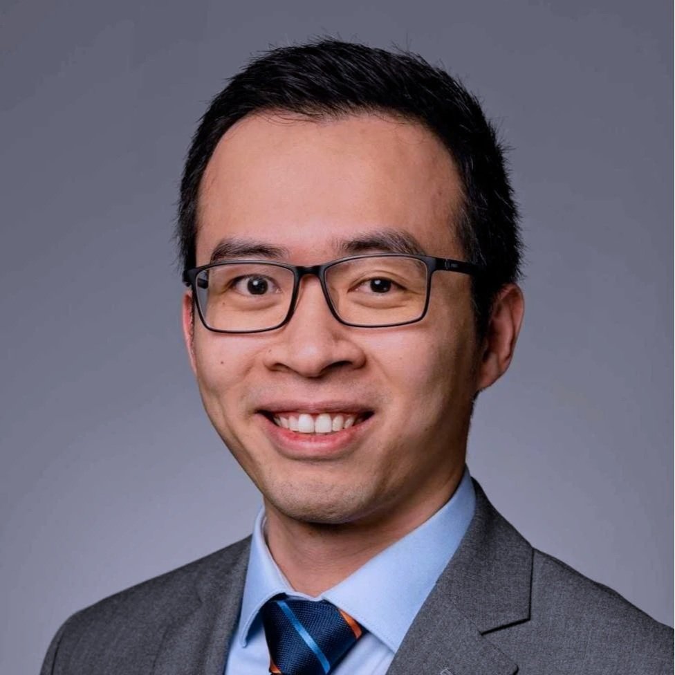 A man wearing glasses, a gray suit, light blue shirt, and a striped tie, smiling at the camera against a plain gray background.