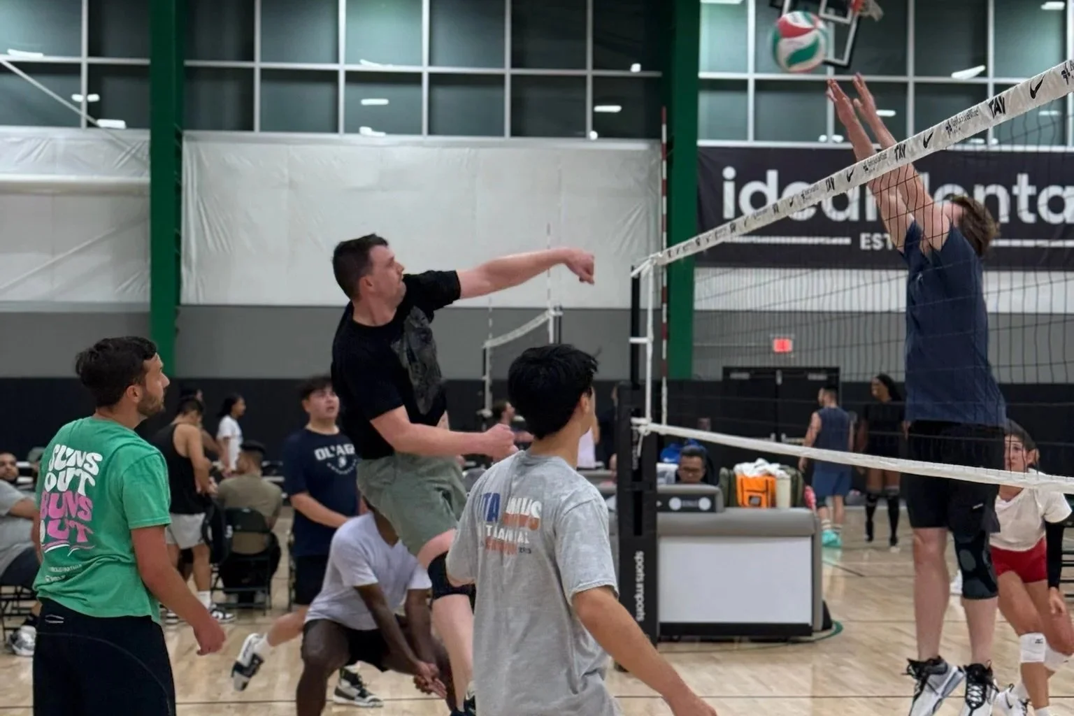 Indoor volleyball game in progress with players jumping and reaching for the ball at the net, while others watch and prepare on the court.