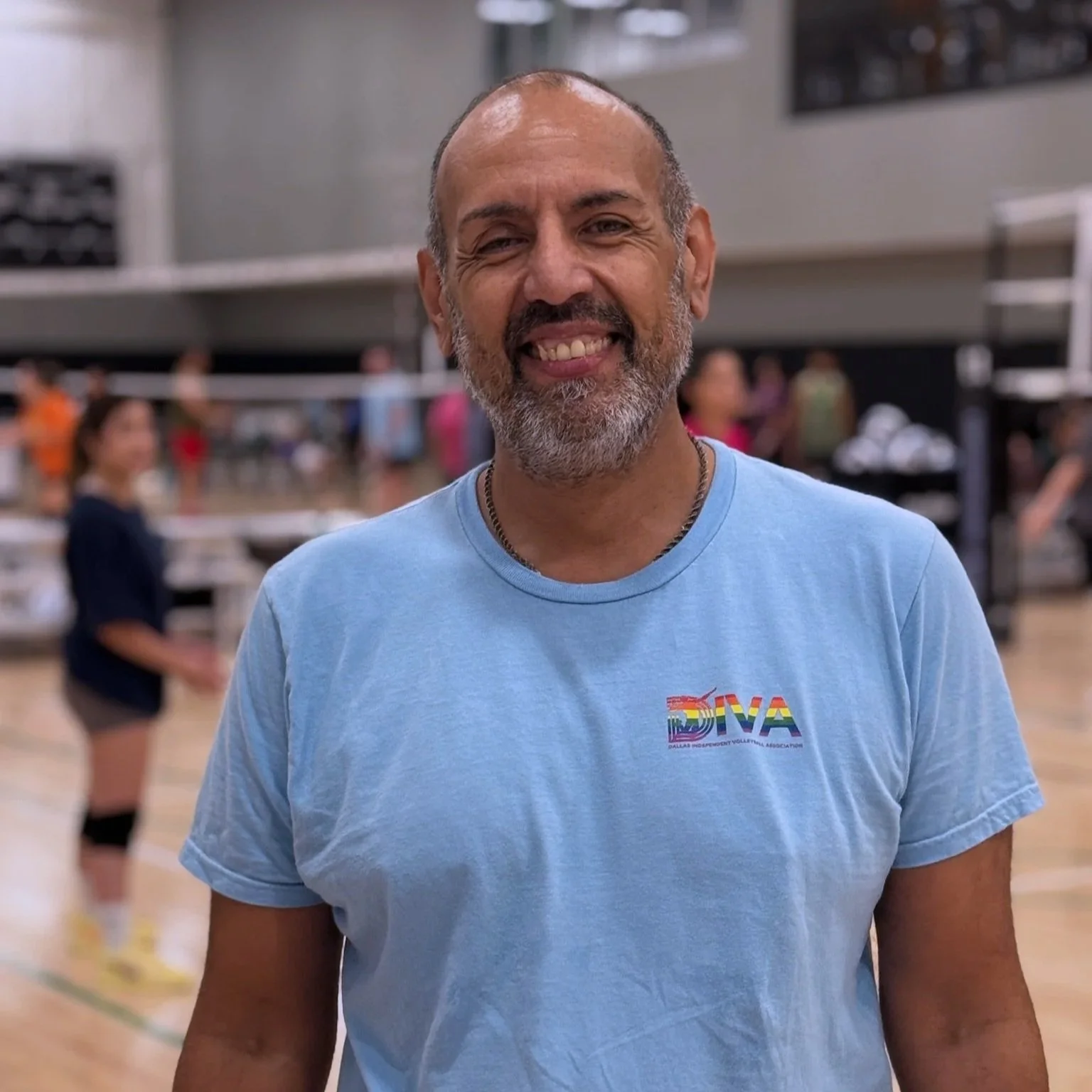 A smiling man with a beard and bald head wearing a light blue t-shirt with rainbow-colored text and a volleyball graphic standing in an indoor volleyball court.