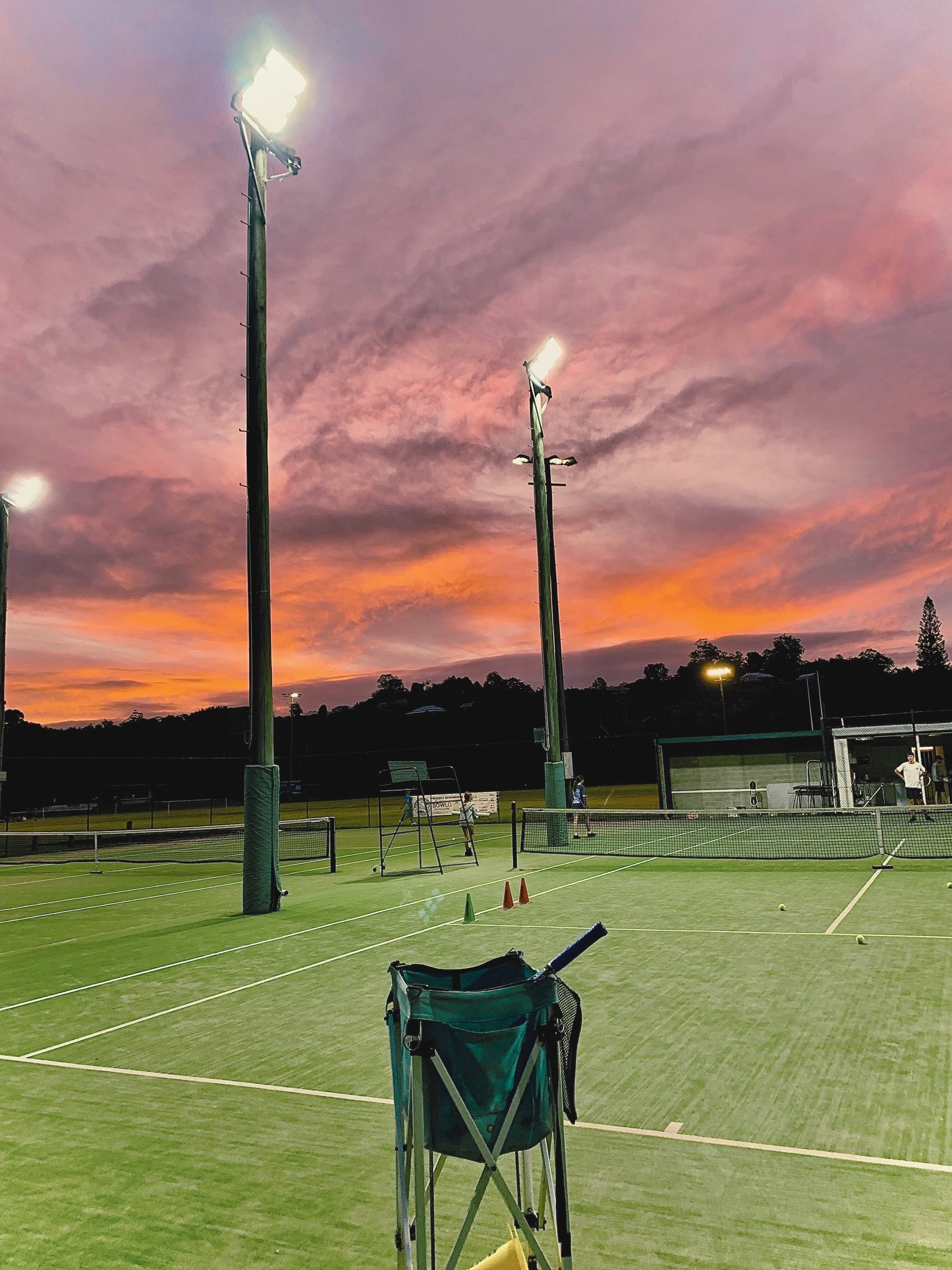 Tennis court at sunset with floodlights, tennis equipment, and a colorful sky.