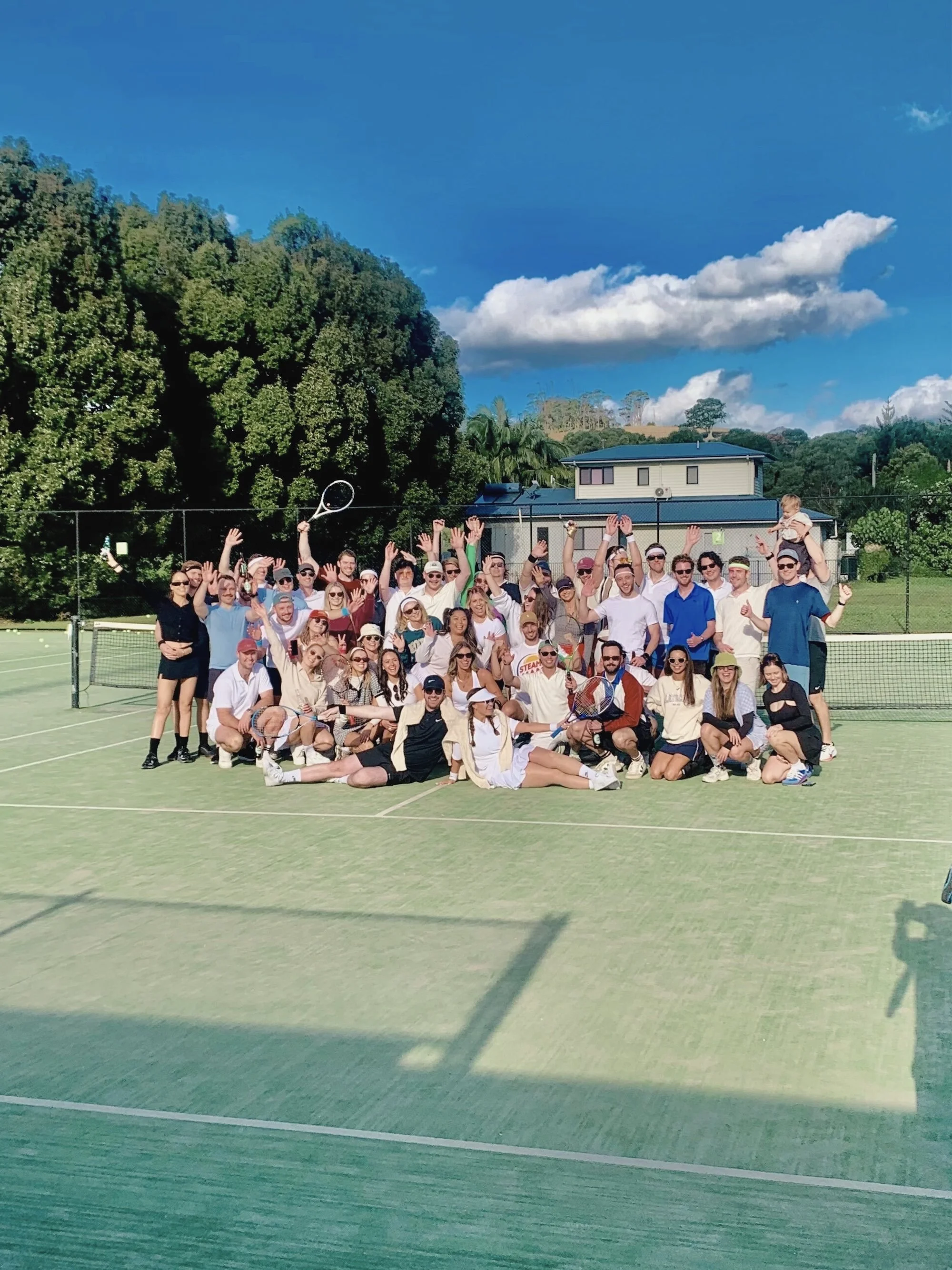 A group of people posing on a tennis court, surrounded by trees and a house, under a blue sky with clouds. Many individuals are smiling and raising their hands in celebration.