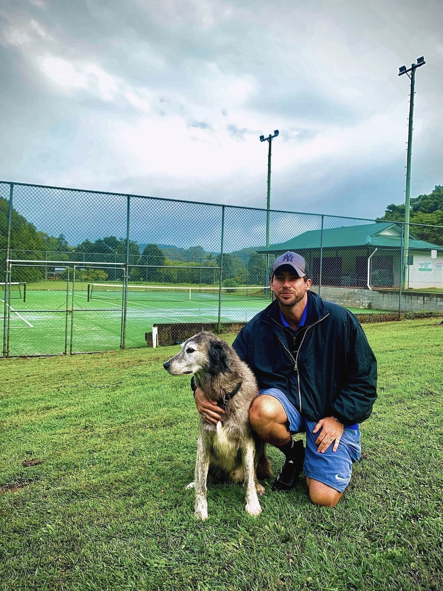 Man kneeling next to a large dog on a grassy field, near a tennis court with fencing in the background. The sky is cloudy, and the man is wearing a baseball cap, jacket, and shorts.