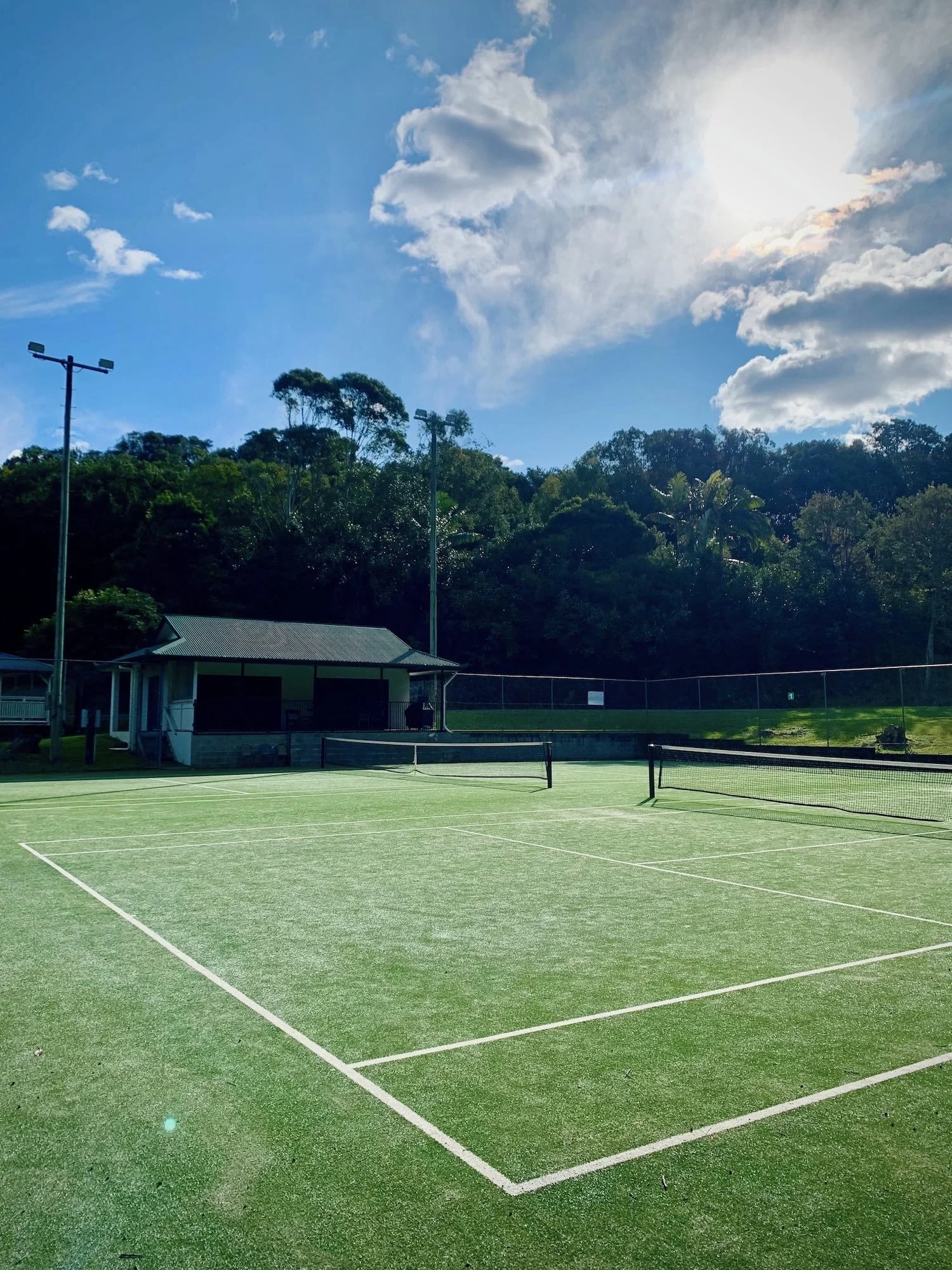 Outdoor tennis court with artificial grass, surrounded by trees and a building under a blue sky with sun and clouds.