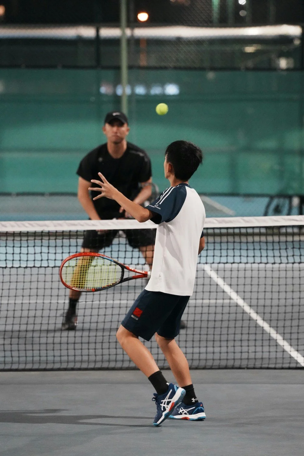 Two people playing tennis on a court, focusing on the player in the foreground preparing to hit a ball with a racket. The opponent is visible near the net.