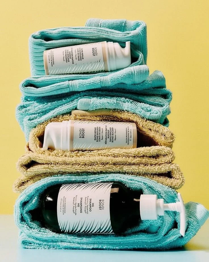 Stacked folded towels with bottles of shower gel, body wash, and shampoo placed on top of and inside the towels against a yellow background.