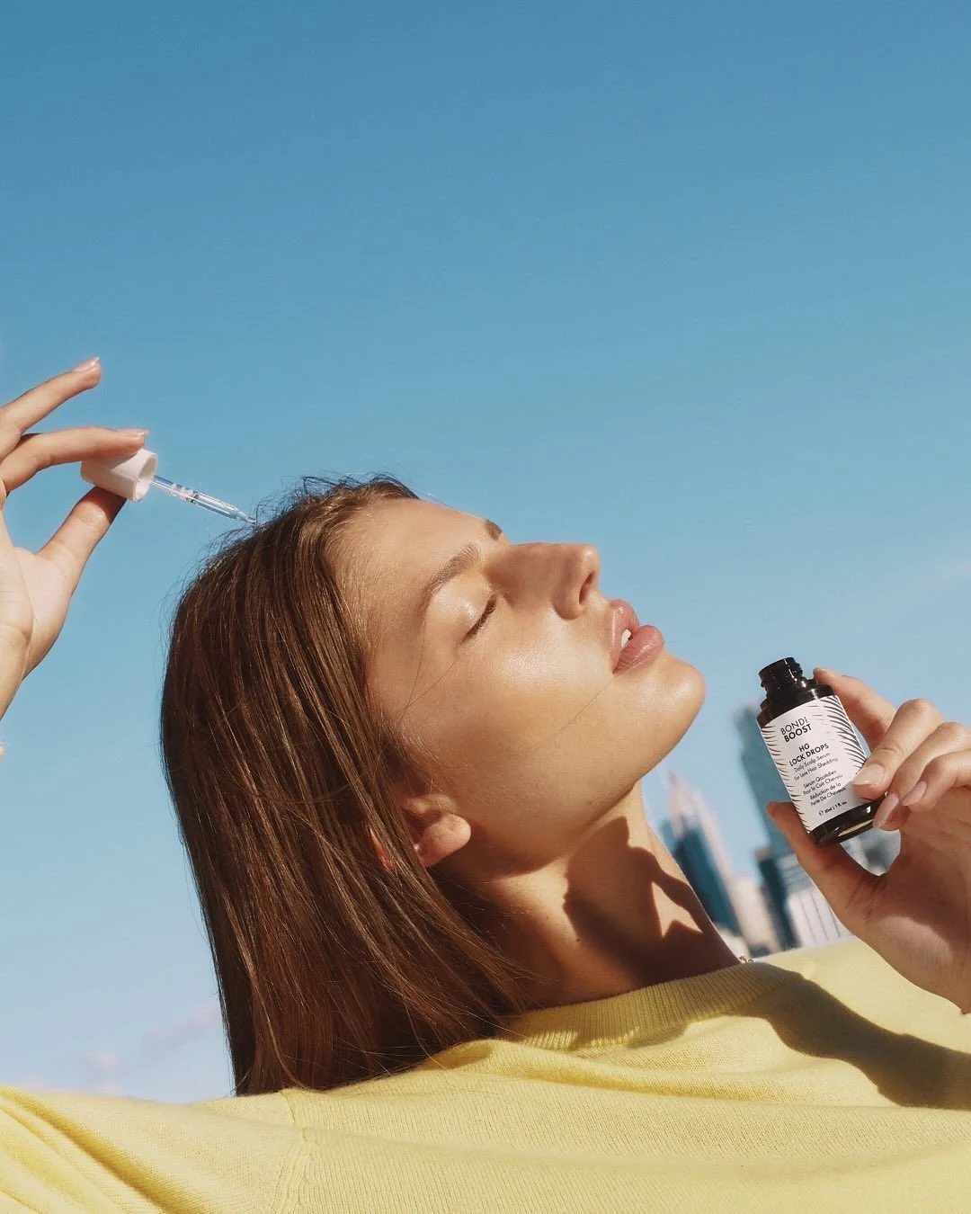 A woman with closed eyes is outdoors, holding a small dark bottle of skincare serum in her left hand and a dropper in her right hand, with a bright blue sky in the background.