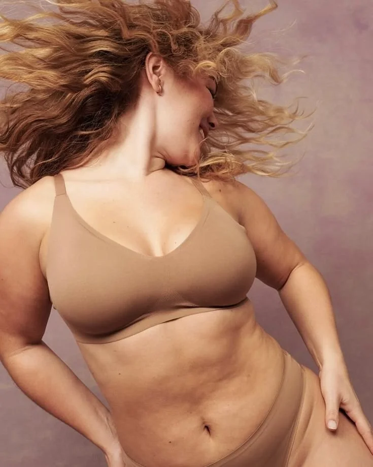 A woman with long curly hair flowing, wearing a beige sports bra and matching bottoms, standing against a pinkish background.