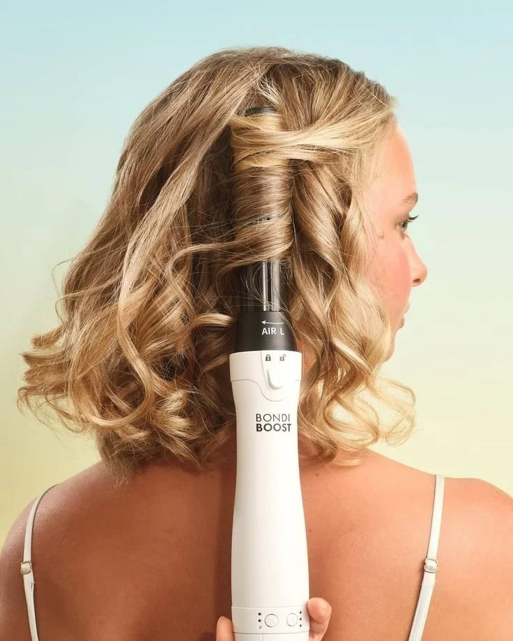 A woman with curly blonde hair is holding a curling iron labeled "Bondi Boost" against her hair.