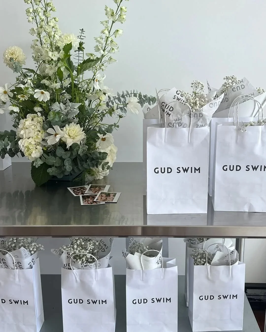 Arrangement of white flowers in a vase, Polaroid photos on a table, and white paper gift bags with the text 'GUD SWIM' on a table in a minimalist setting.