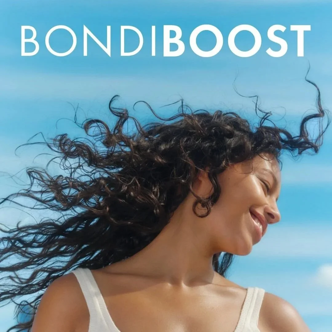 A woman with long, curly hair smiling and enjoying the breeze outdoors with a blue sky in the background, promoting Bondi Boost hair care products.