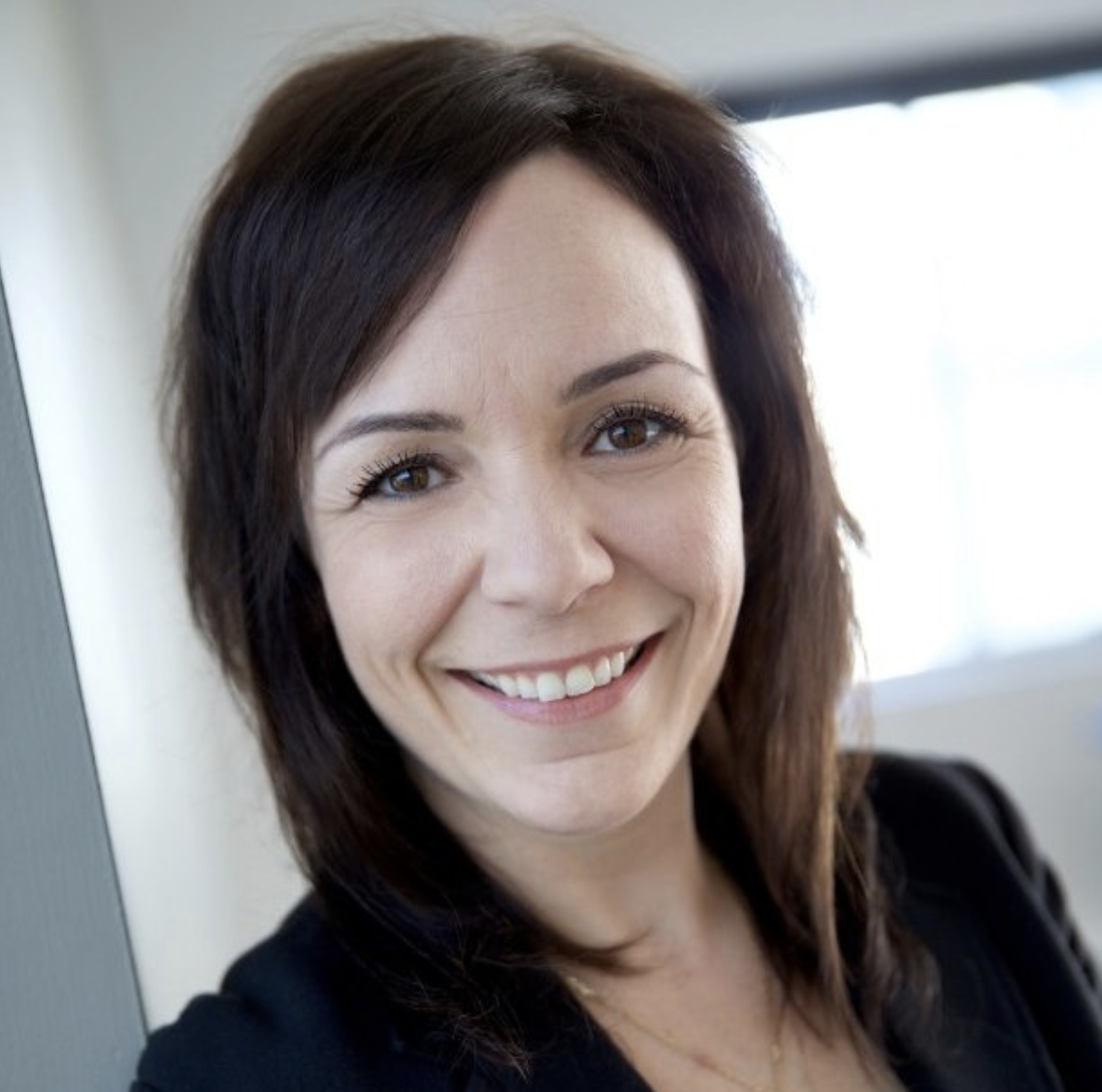 Portrait d'une femme souriante avec des cheveux bruns, portant un blazer noir, dans un environnement intérieur lumineux.