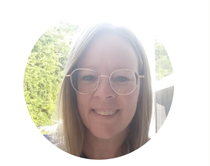 Une femme souriante porte des lunettes et a les cheveux longs, avec un fond lumineux et des arbres verts.