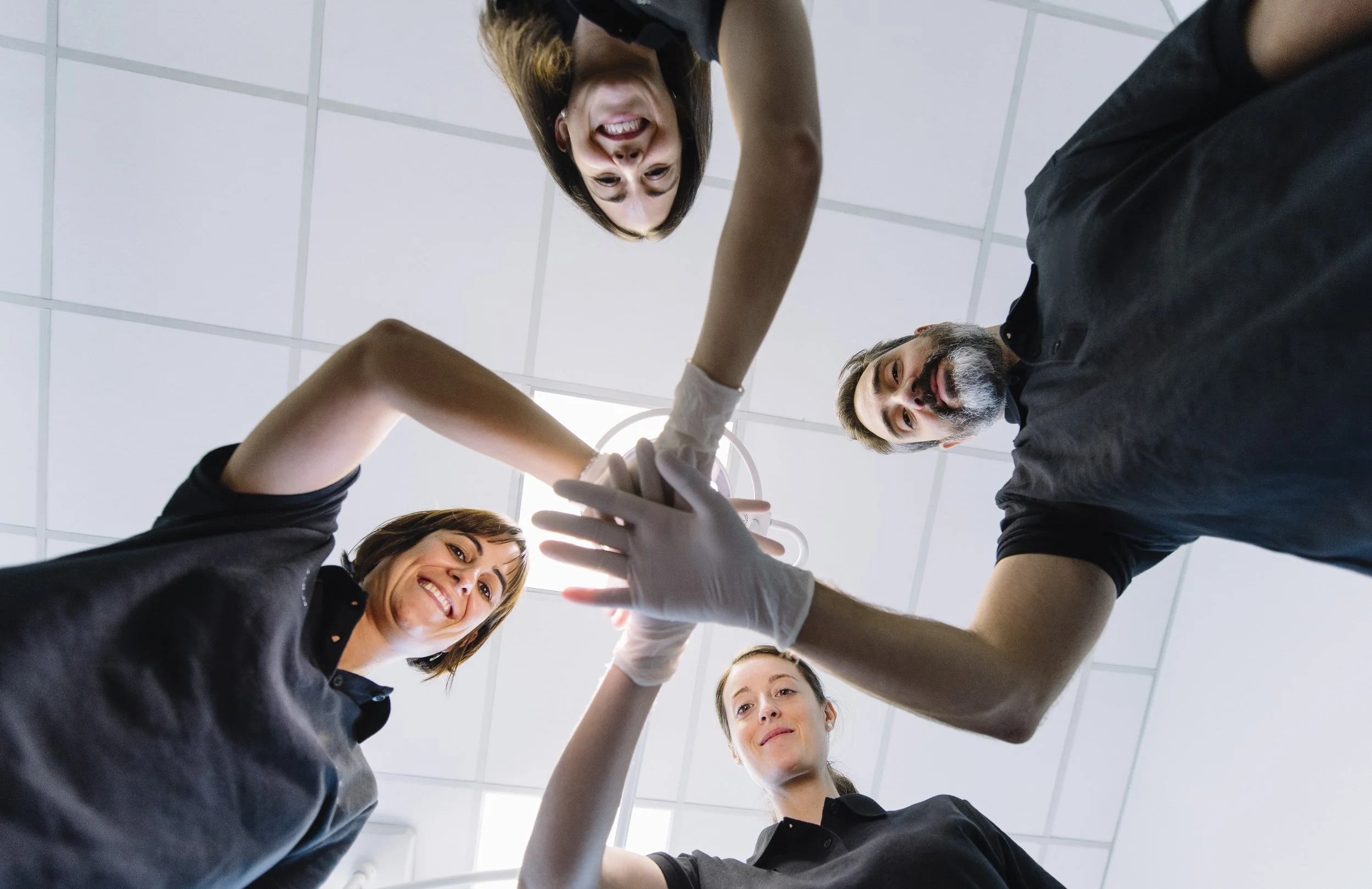 Quatre personnes, deux femmes et deux hommes, se donnent la main en cercle, souriantes en regardant vers le haut dans une salle au plafond blanc.