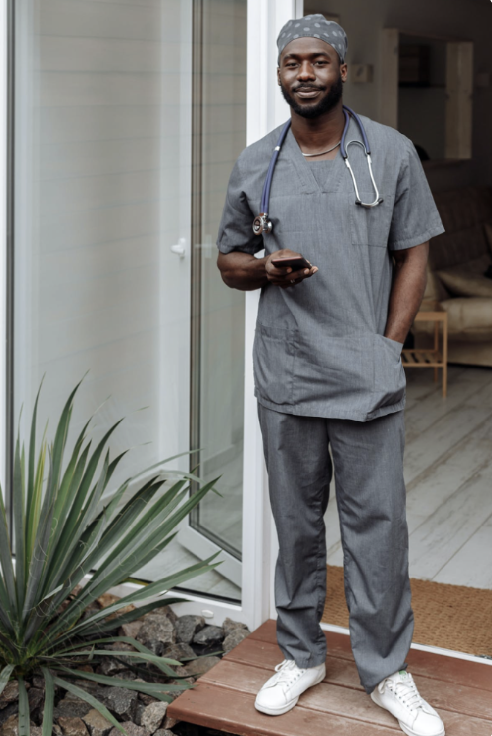 Un professionnel de la santé en uniforme gris avec un stéthoscope autour du cou, tenant un téléphone portable, debout sur une terrasse en bois devant une porte-fenêtre.