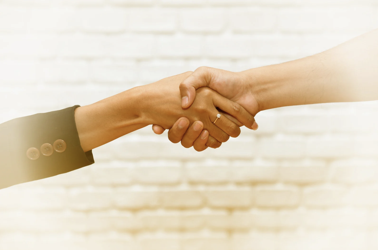 Deux personnes se serrent la main, une personne porte une veste noire avec des boutons beige, l'autre personne porte une peau claire et une alliance. Fond en briques blanches.