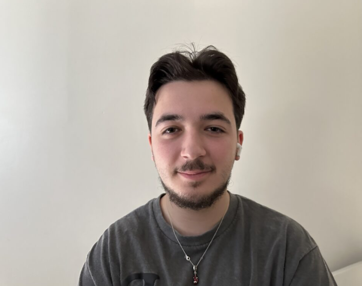 Un jeune homme avec des écouteurs, portant une chaîne, souriant devant un mur beige clair.
