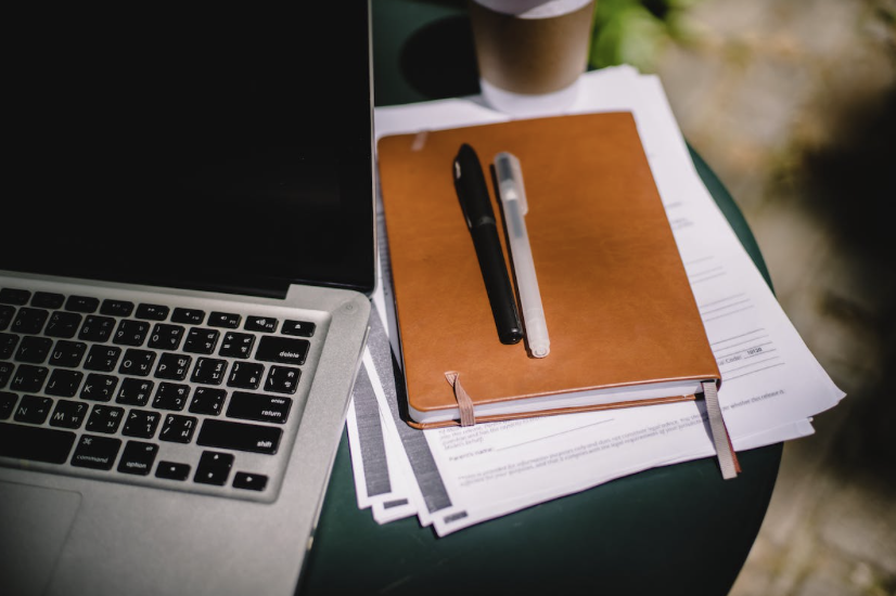 Un ordinateur portable à côté d'un carnet orange, deux stylos, et des papiers sur une table ronde en extérieur.