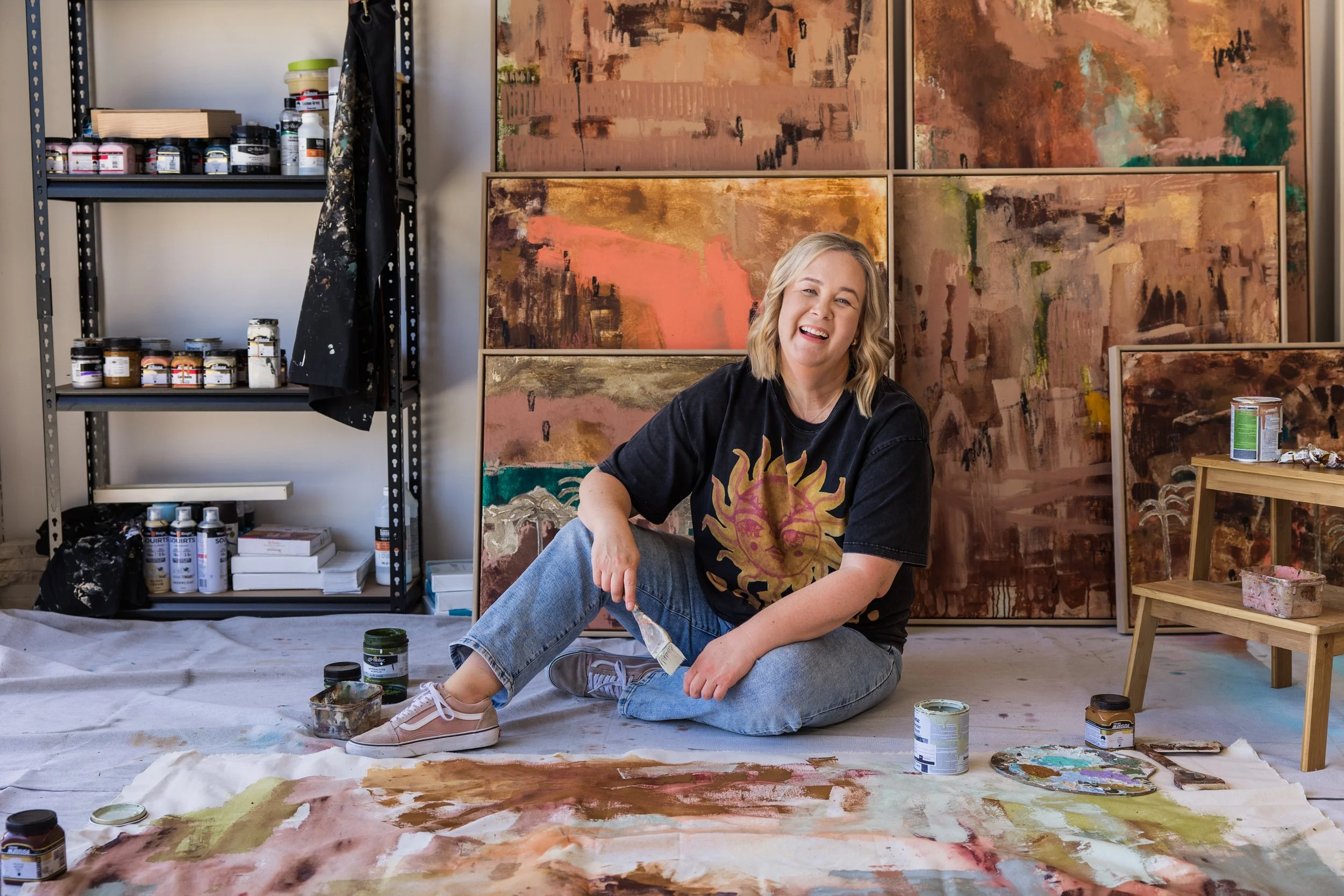 A woman sitting on the floor of an art studio, surrounded by abstract paintings and art supplies, smiling while holding a paintbrush.