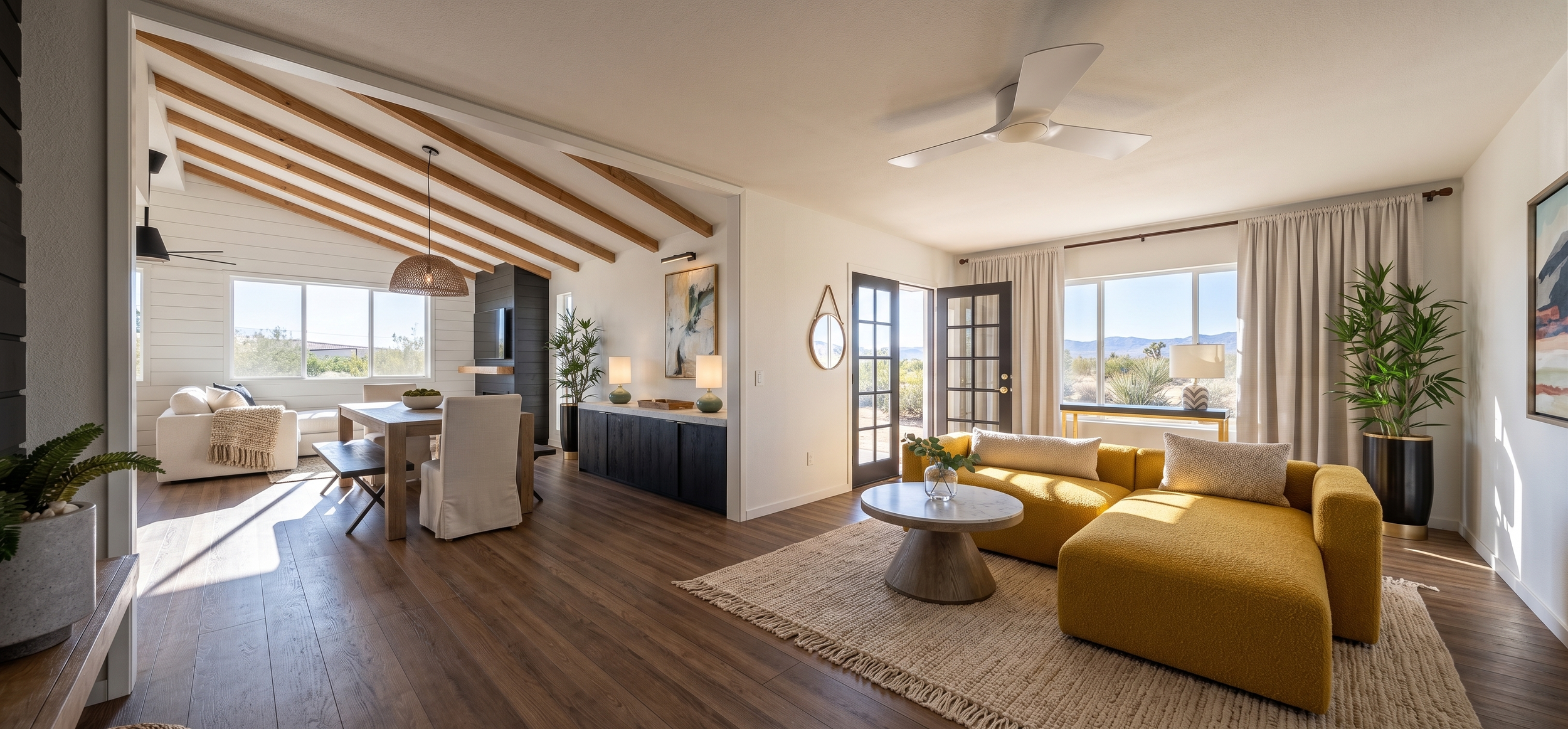 Open-concept living and dining area with large windows, a yellow sofa, a round coffee table, and a wooden dining table with white chairs, featuring wood ceiling beams and desert landscape views outside.