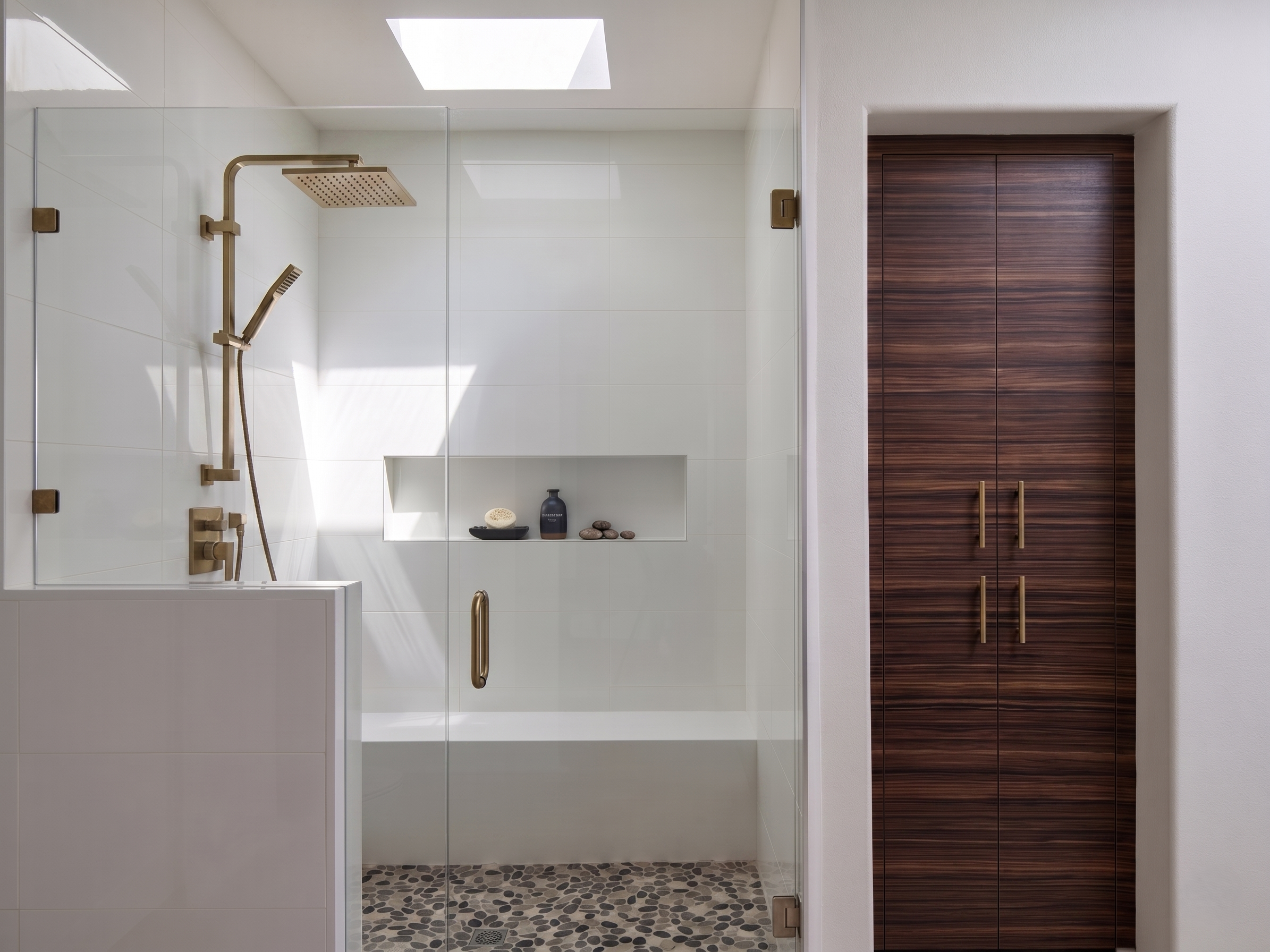 Modern walk-in shower with glass door, white tiled walls, a rain shower head, handheld shower, built-in niche with soap and shampoo, pebble tile floor, and a wooden cabinet outside the shower.