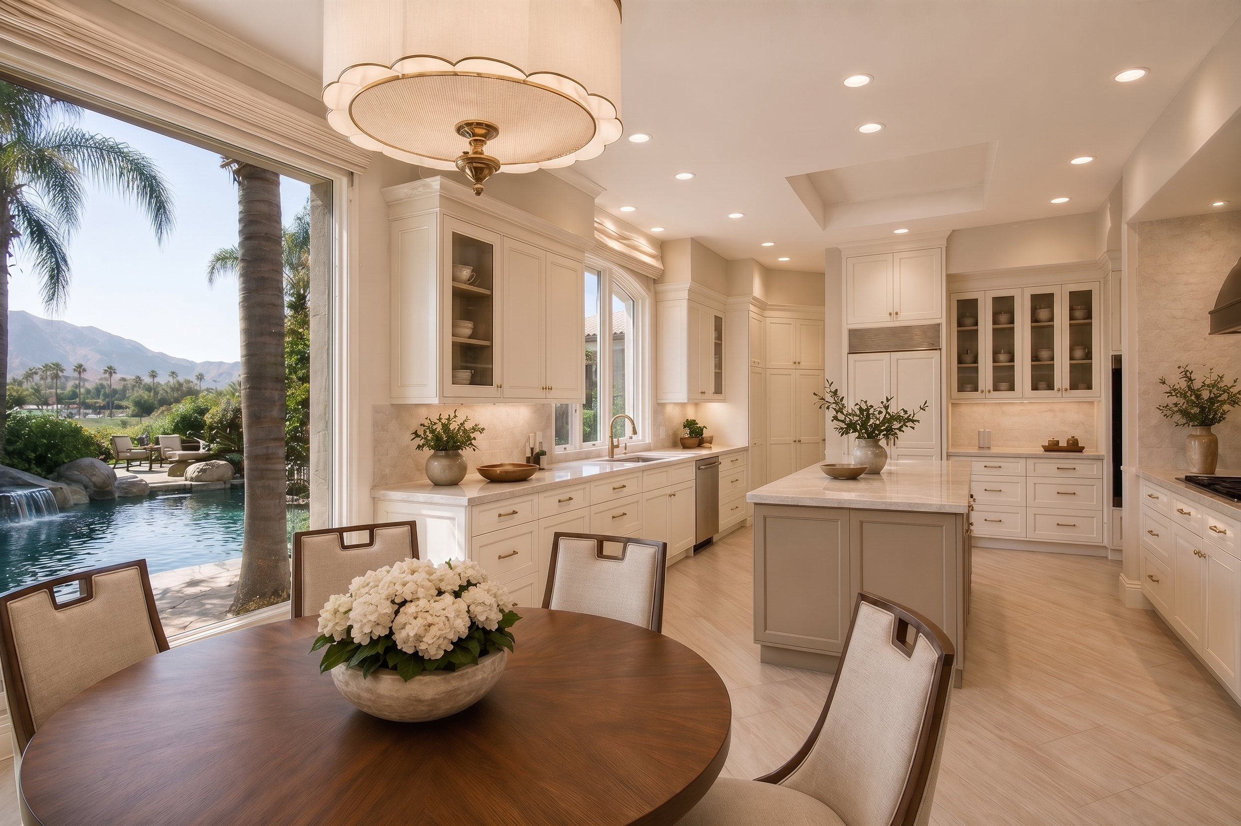 Bright kitchen with white cabinets, marble countertops, and large windows overlooking a backyard with a pool, palm trees, and mountains in the distance.