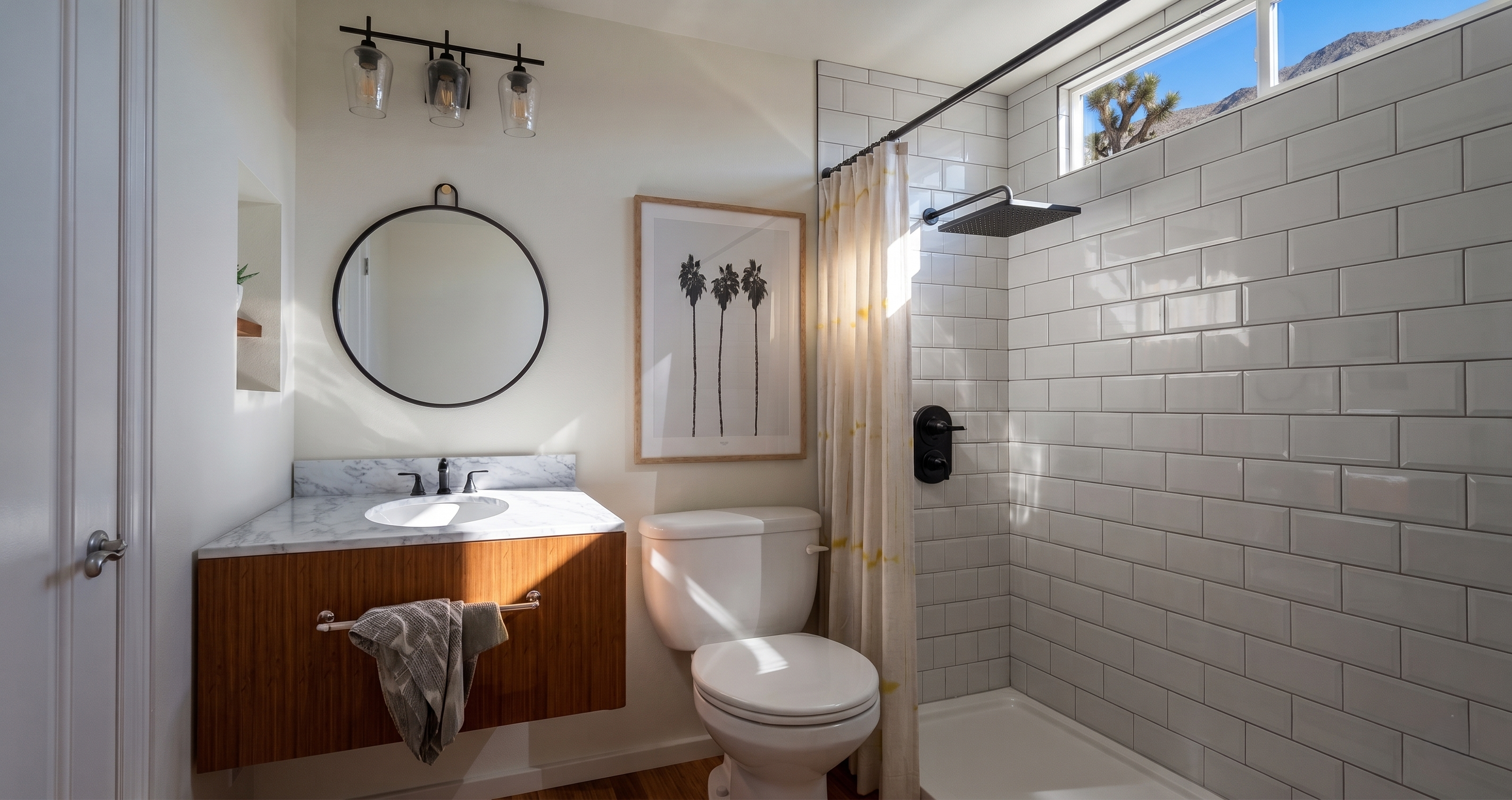 A modern bathroom featuring a white tile shower with a rainfall showerhead, a small vanity with a marble countertop, a round mirror, a framed picture of three palm trees, a toilet, a towel rail with a towel, and a large window showing a mountain view