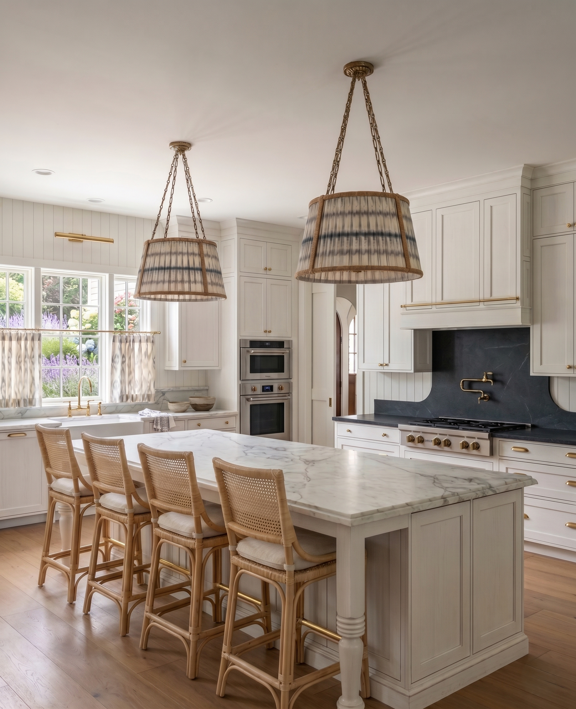 A kitchen with white cabinetry, a marble island with a white base, and a row of six rattan chairs with white cushions arranged around the island. Two large hanging lamps with striped shades are suspended from the ceiling over the island. The background features a window with a view of a garden with purple and white flowers, and built-in appliances.