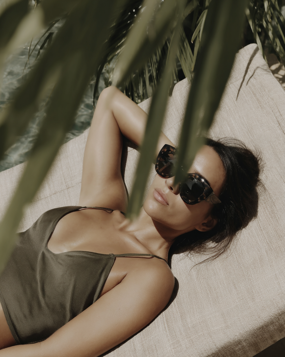 A woman in sunglasses and a dark green swimsuit relaxing on a cushioned lounge chair outdoors, with palm leaves partially covering her and a body of water in the background.