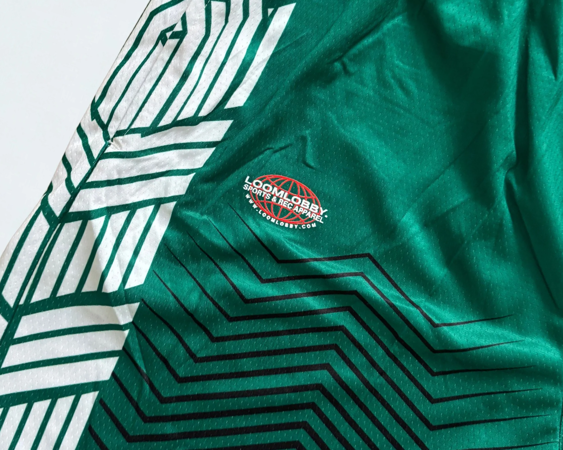 Close-up of a green sports jersey with black and white geometric patterns and a logo that says 'LOOMLOBY SPORTS & APPAREL' with a red globe symbol.