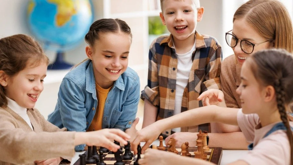 Діти та жінка грають у шахи за столом, посміхаються, на задньому плані — глобус.