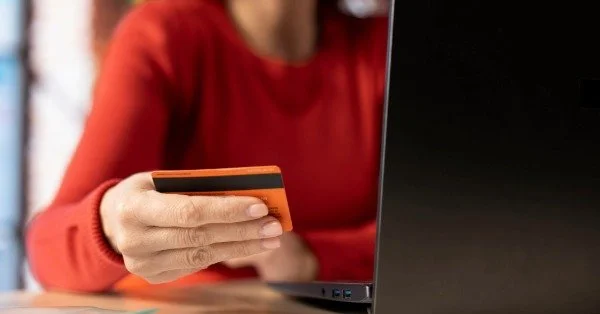 Людина в червоній сорочці тримає кредитну картку біля ноутбука.