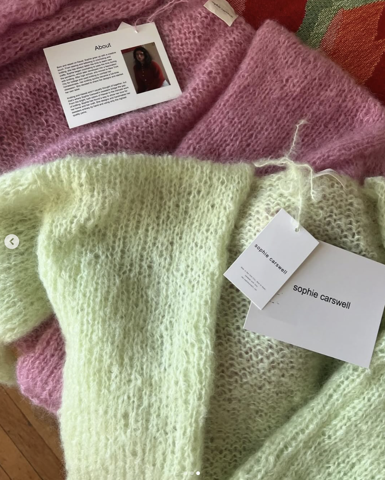Several knit sweaters in pink and light yellow colors displayed with tags from the designer Sophie Carswell and one tag providing an 'About' description and a photo of a woman, likely the designer.