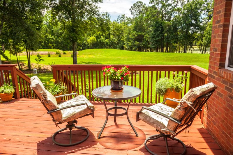 Wooden deck with two cushioned chairs and a round glass table, potted flowers, and a view of a green lawn and trees.