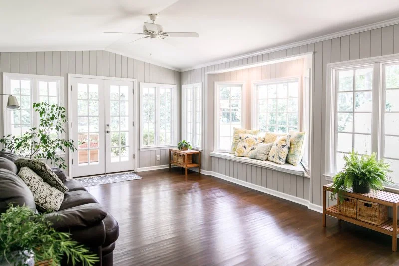 Bright living room with hardwood floors, a brown sofa, French doors, large windows with a cushioned window seat, and indoor plants.