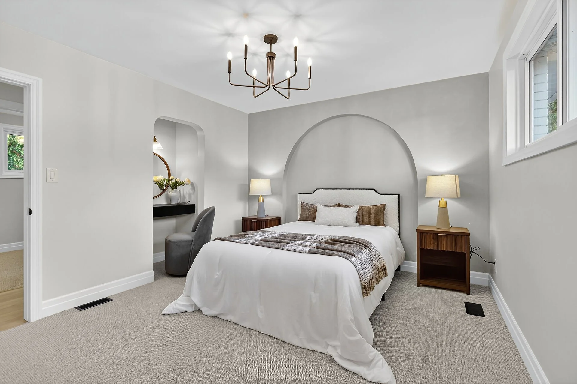 A modern bedroom with a white bed, beige headboard, brown and white pillows, and a striped blanket. Two wooden nightstands with lamps flank the bed. A ceiling chandelier with multiple candle-like bulbs hangs above. There is a small vanity with a mirror and flowers on the left, and two windows on the right allow natural light.