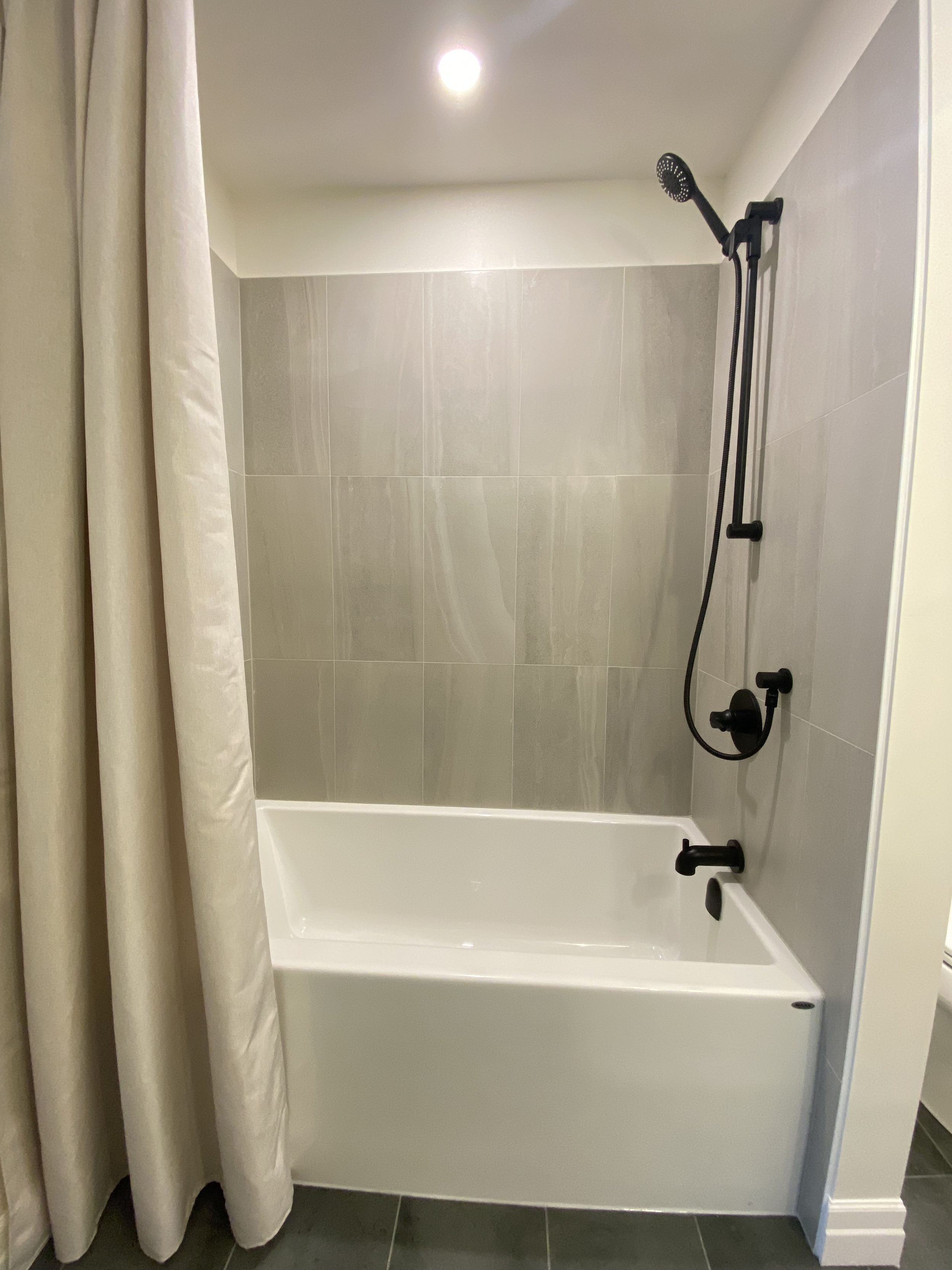 Bathroom shower with grey tiled walls, a black showerhead, and a white bathtub. There is a beige curtain on the left, and the floor is dark grey tiles.