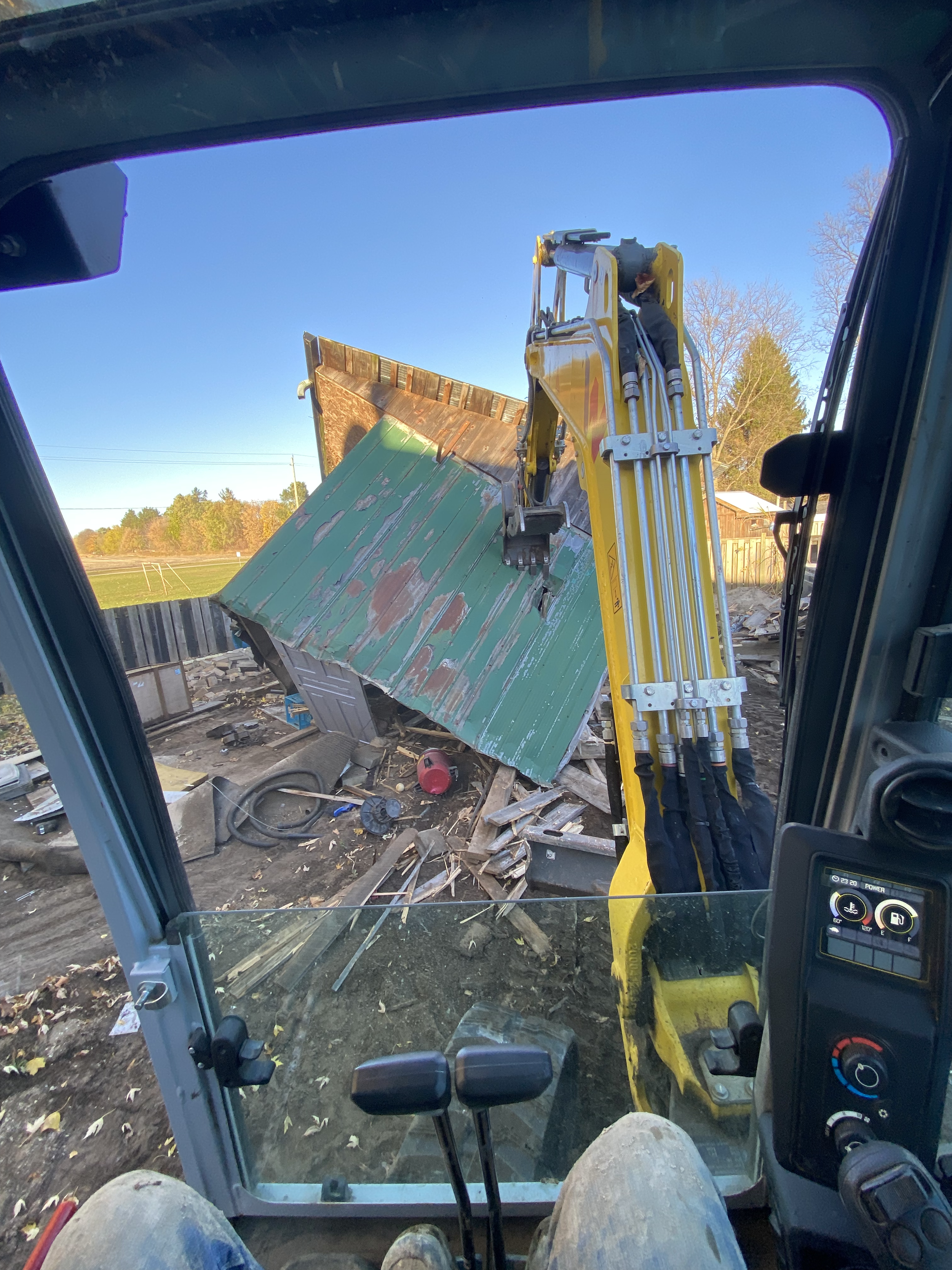 View from inside a construction vehicle's cabin showing a worker operating an excavator with a yellow arm, demolishing a small green metal shed with a rusted roof, surrounded by debris and construction tools.
