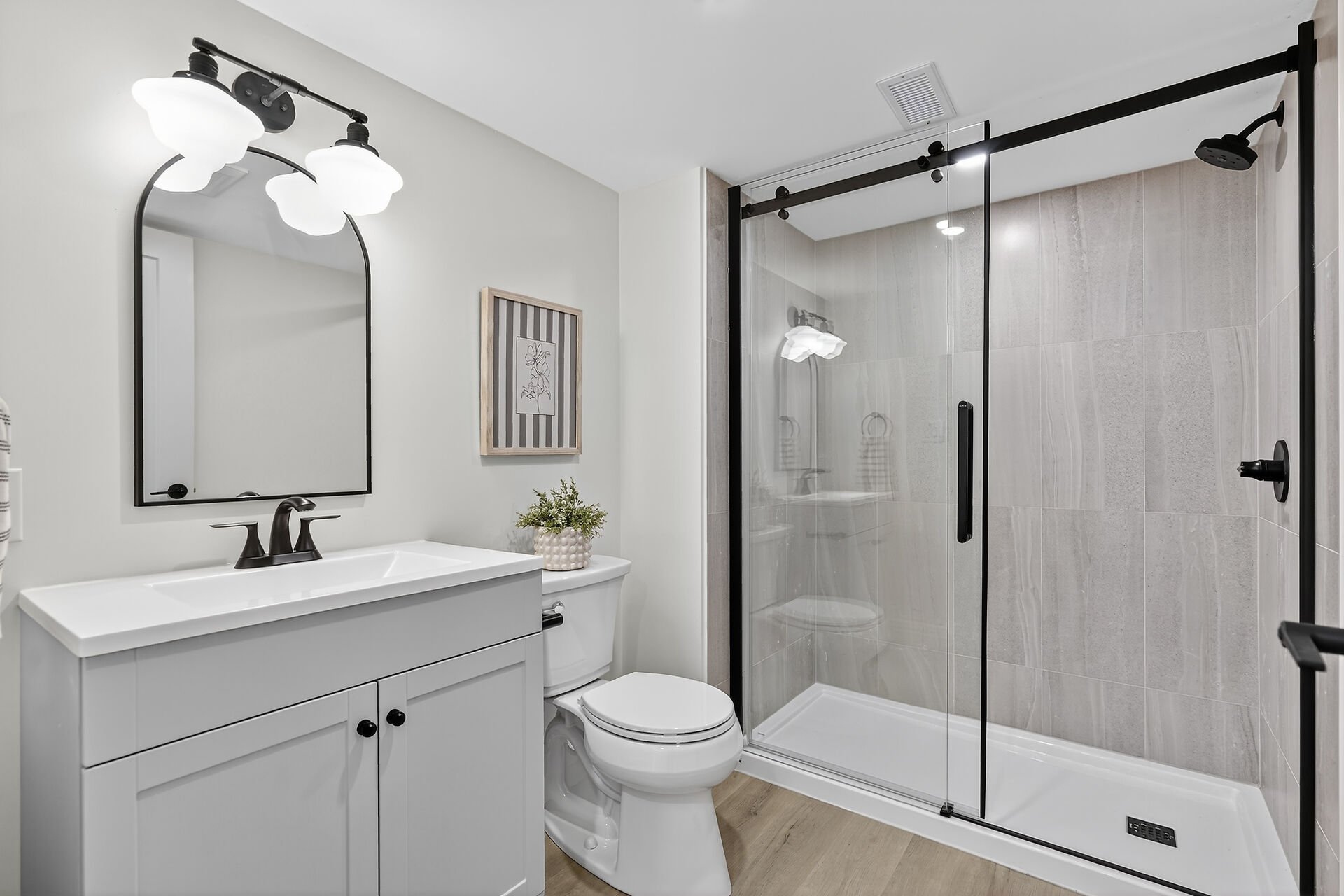 Modern bathroom with white vanity, black fixtures, wall mirror, toilet, framed artwork, potted plant, and a glass-enclosed shower with marble tiles.