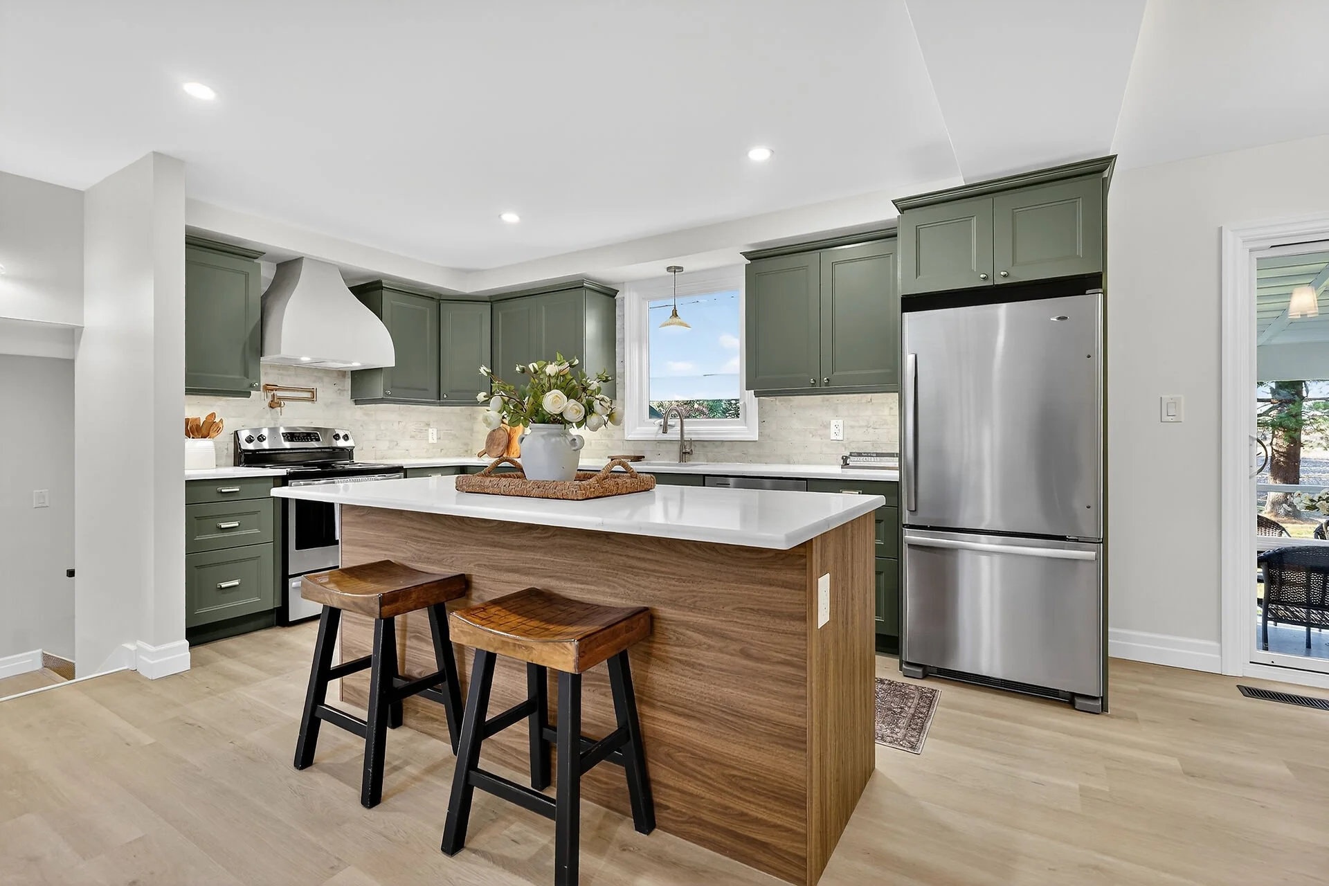 Modern kitchen with green cabinets, stainless steel refrigerator, island with white countertop, two wooden stools, and a flower arrangement in a vase.