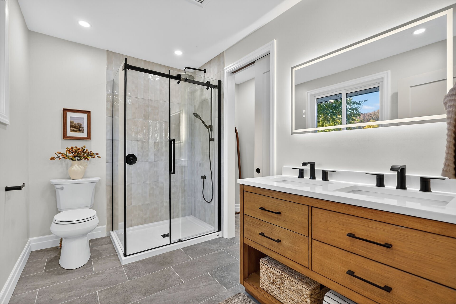 Modern bathroom with a walk-in shower with black framing and a glass door, a white toilet with a vase of flowers on top, a wooden vanity with a white countertop and black fixtures, and a large mirror above the vanity. There's a window revealing trees outside.