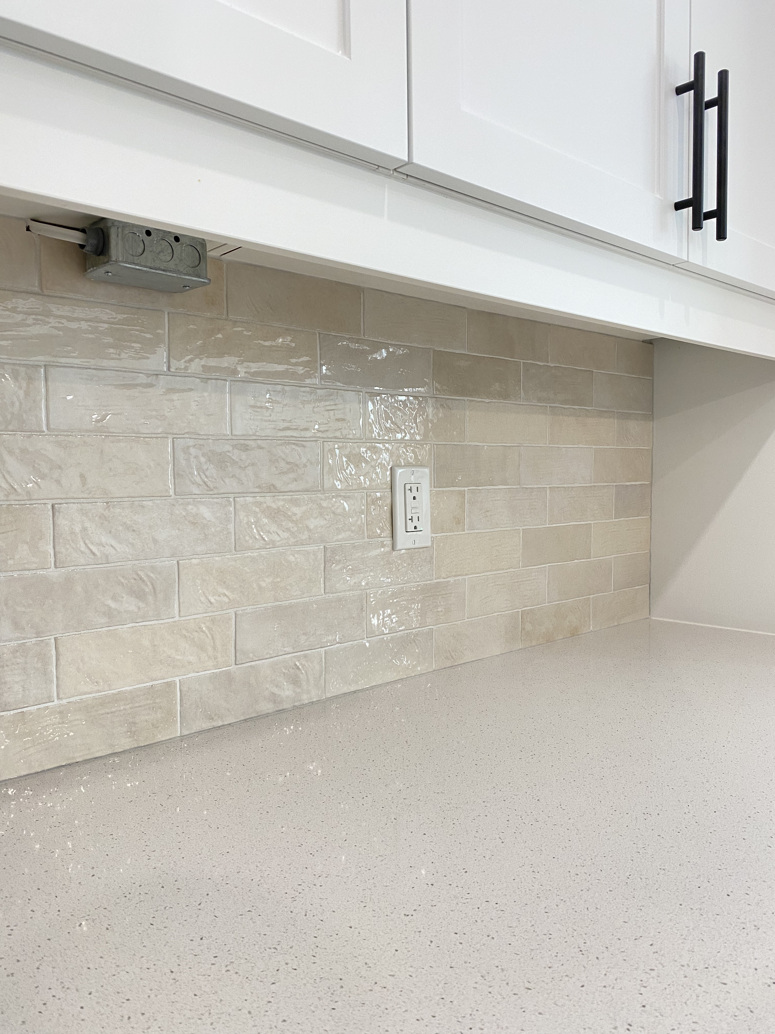 Kitchen backsplash with beige subway tiles, an electrical outlet, and part of a white cabinet with black handles.