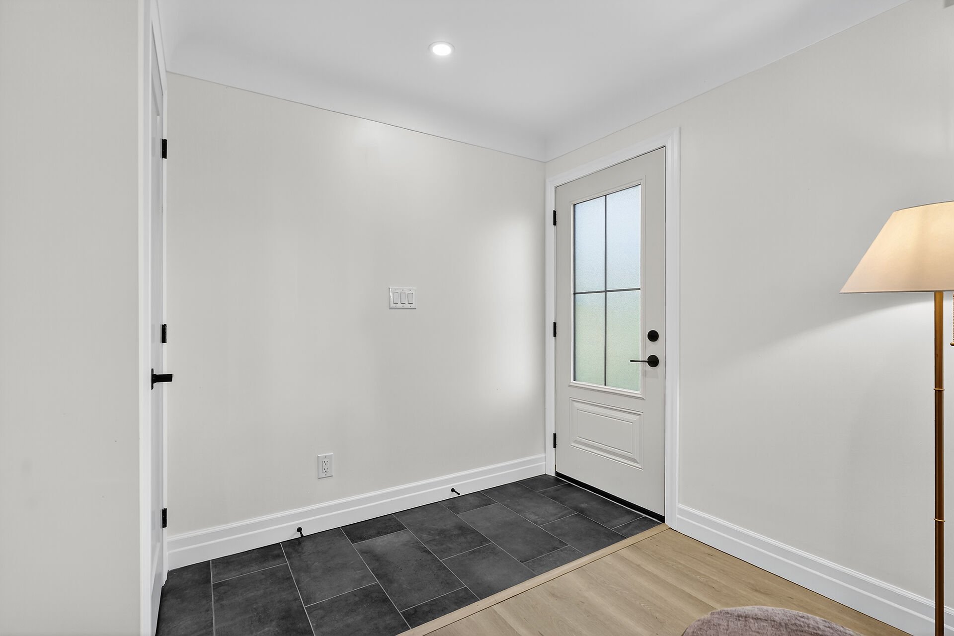 Empty entryway with black tiled floor, white walls, white door with window, and a beige floor lamp.