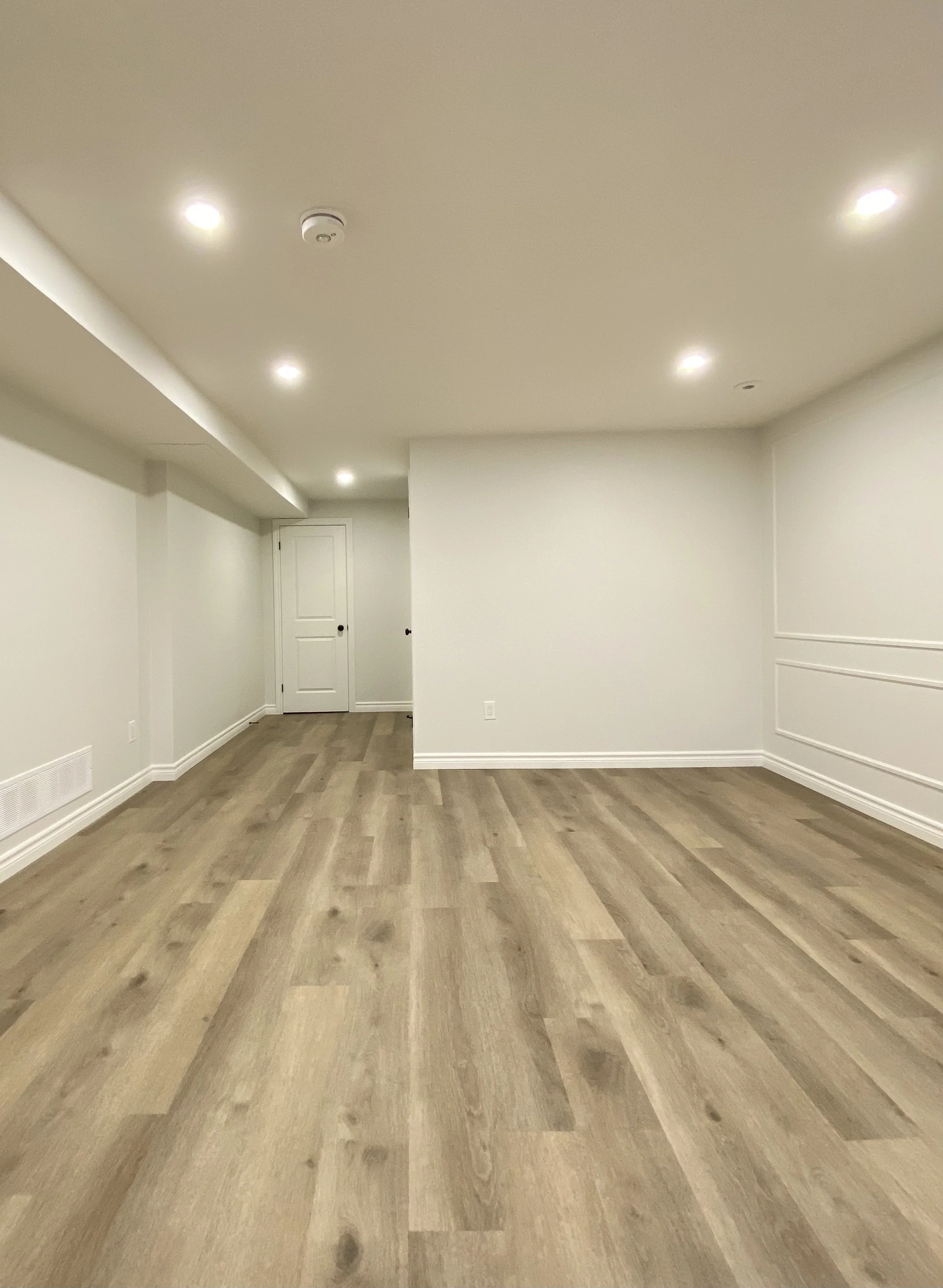 Empty room with wooden flooring, white walls, recessed ceiling lights, and a closed door at the back.