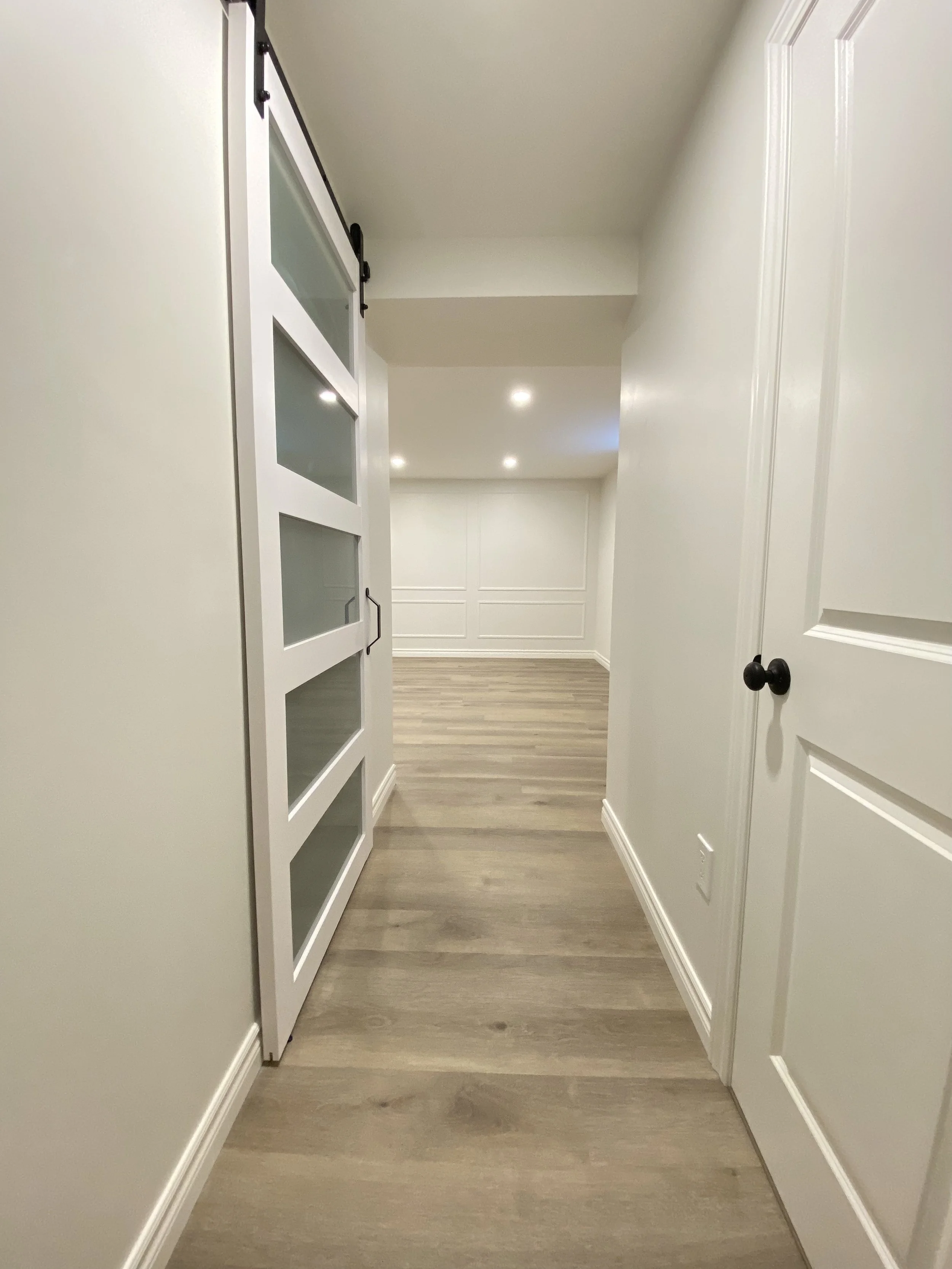 A hallway with white walls, wooden flooring, a door on the right, and a white sliding door with glass panels on the left leading into another room.