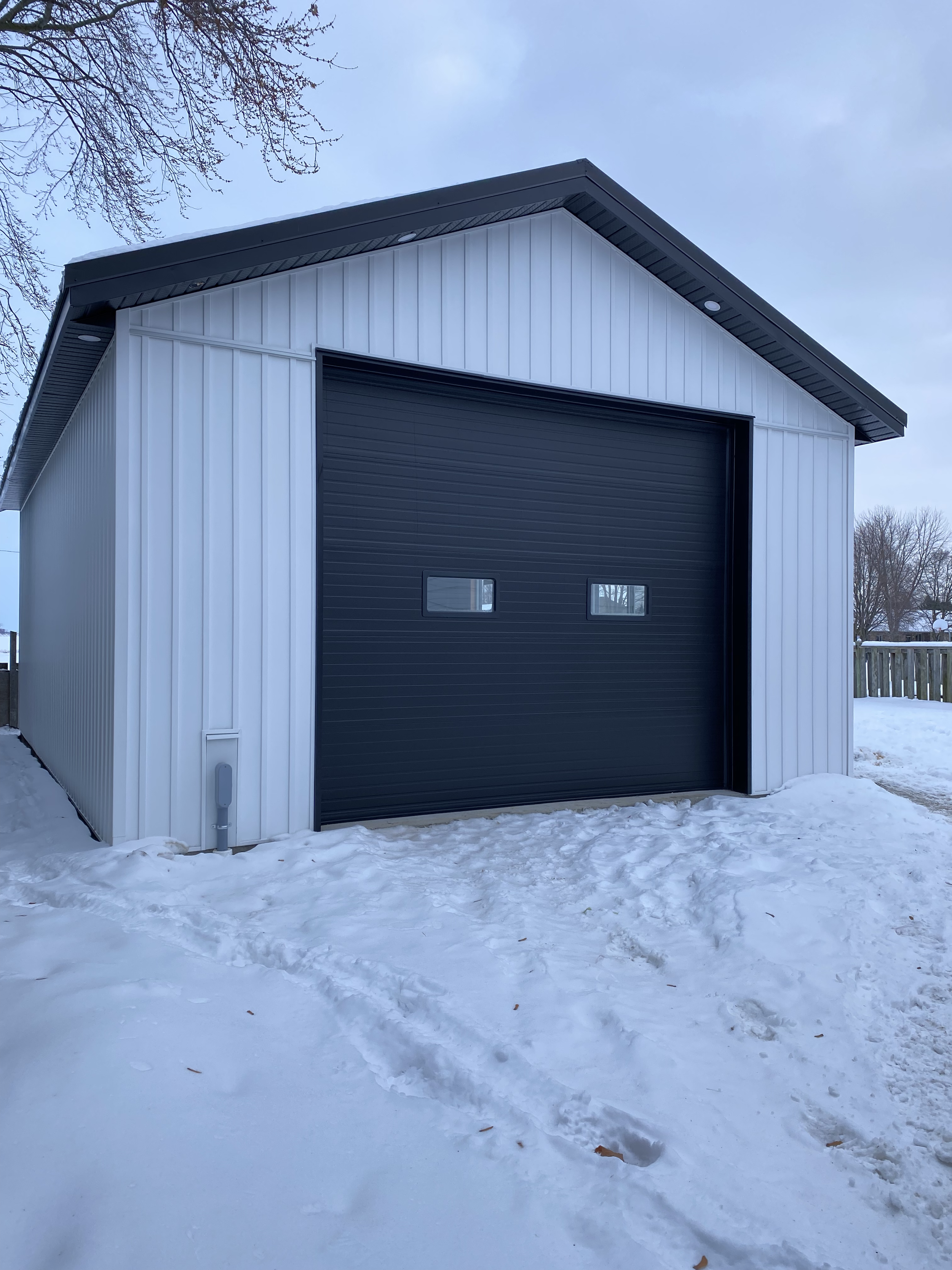 A metal garage building with a black roll-up door and small windows, surrounded by snow in a residential area with trees in the background.