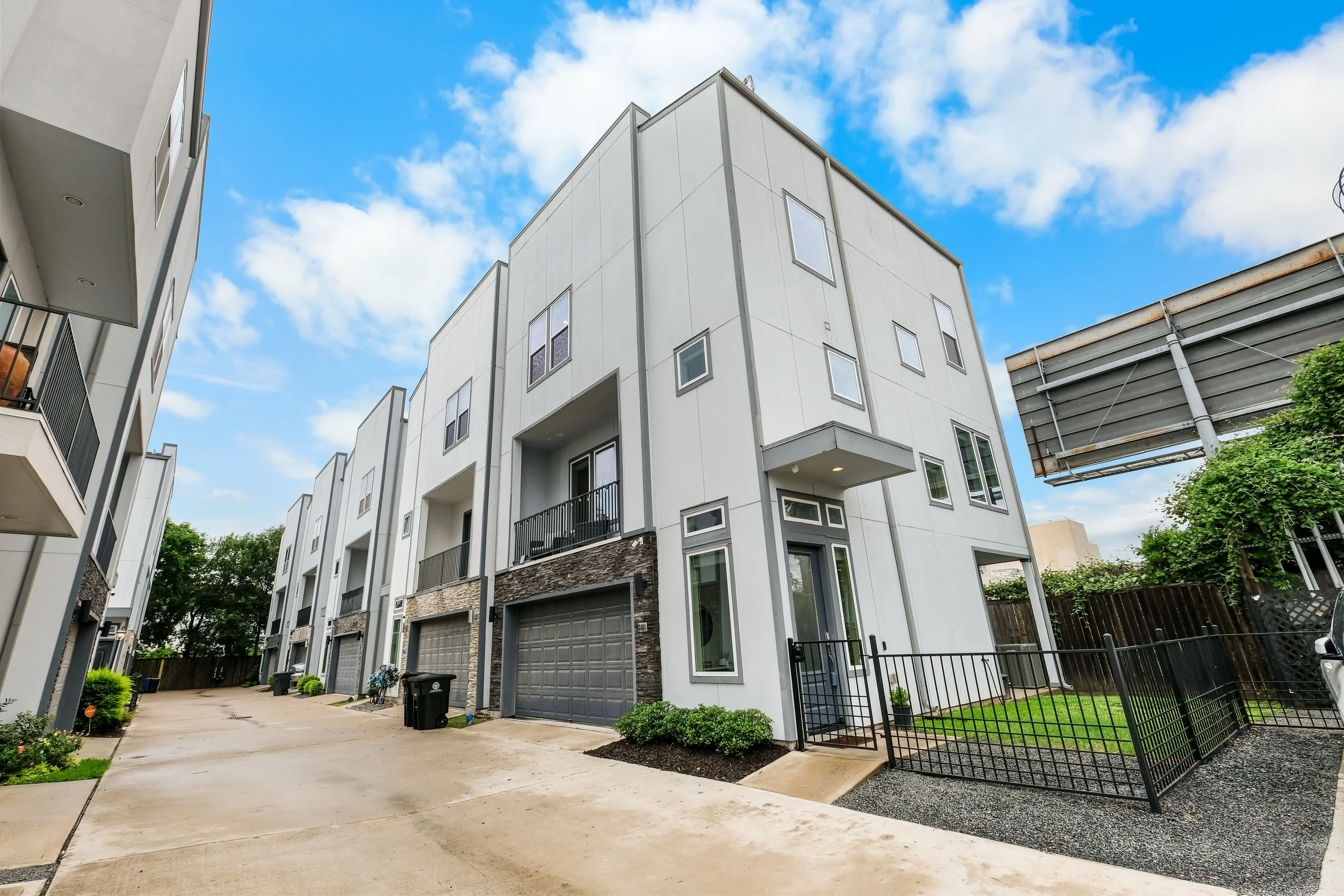 Modern multi-story residential buildings with garage doors and small yards under a partly cloudy sky.