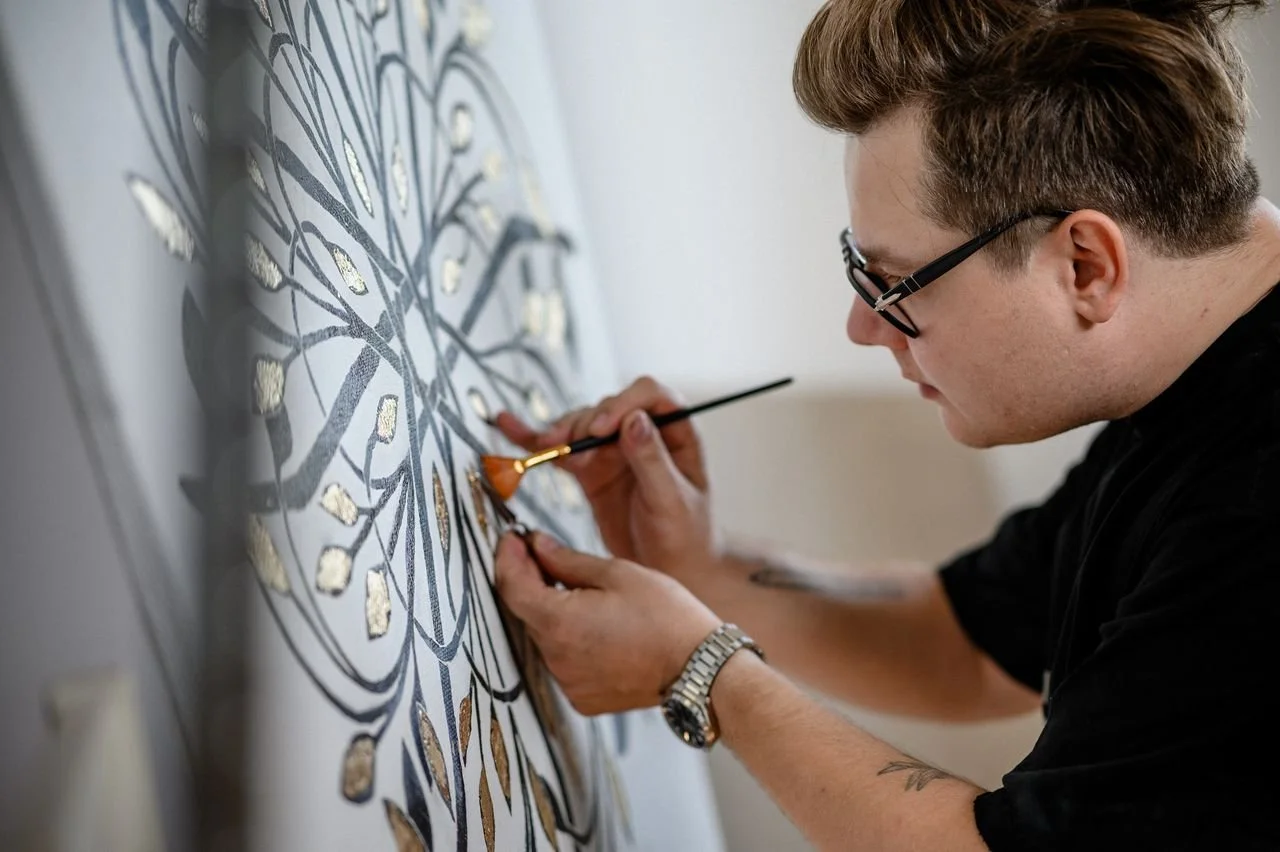 A man wearing glasses, black t-shirt and a watch is working on a large, intricate painting using gold leaf.